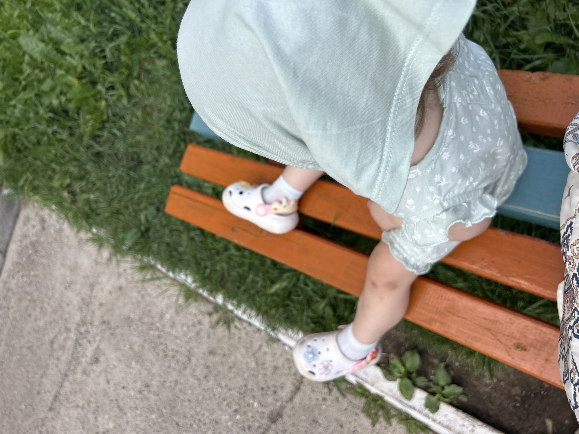 The image shows a close-up view of a young child, likely a toddler, sitting on a wooden park bench. The child is wearing a light green sun hat and a matching light green romper with a subtle floral pattern. White socks and white Crocs, adorned with colorful Jibbitz charms, cover the child's feet. One of the child's legs is bent, and a small bruise is visible on the knee. The bench has orange wooden slats and a blue metal frame, and it is positioned outdoors, with green grass and a concrete pathway visible in the background. Small green plants are growing in the dirt beneath the bench. A patterned fabric, possibly part of another person or an item of clothing, is visible on the right side of the frame. The lighting suggests it is daytime, likely sunny, given the bright exposure and vibrant colors of the scene. The overall impression is a casual outdoor moment, possibly in a park or garden, in Noviy, Russia.