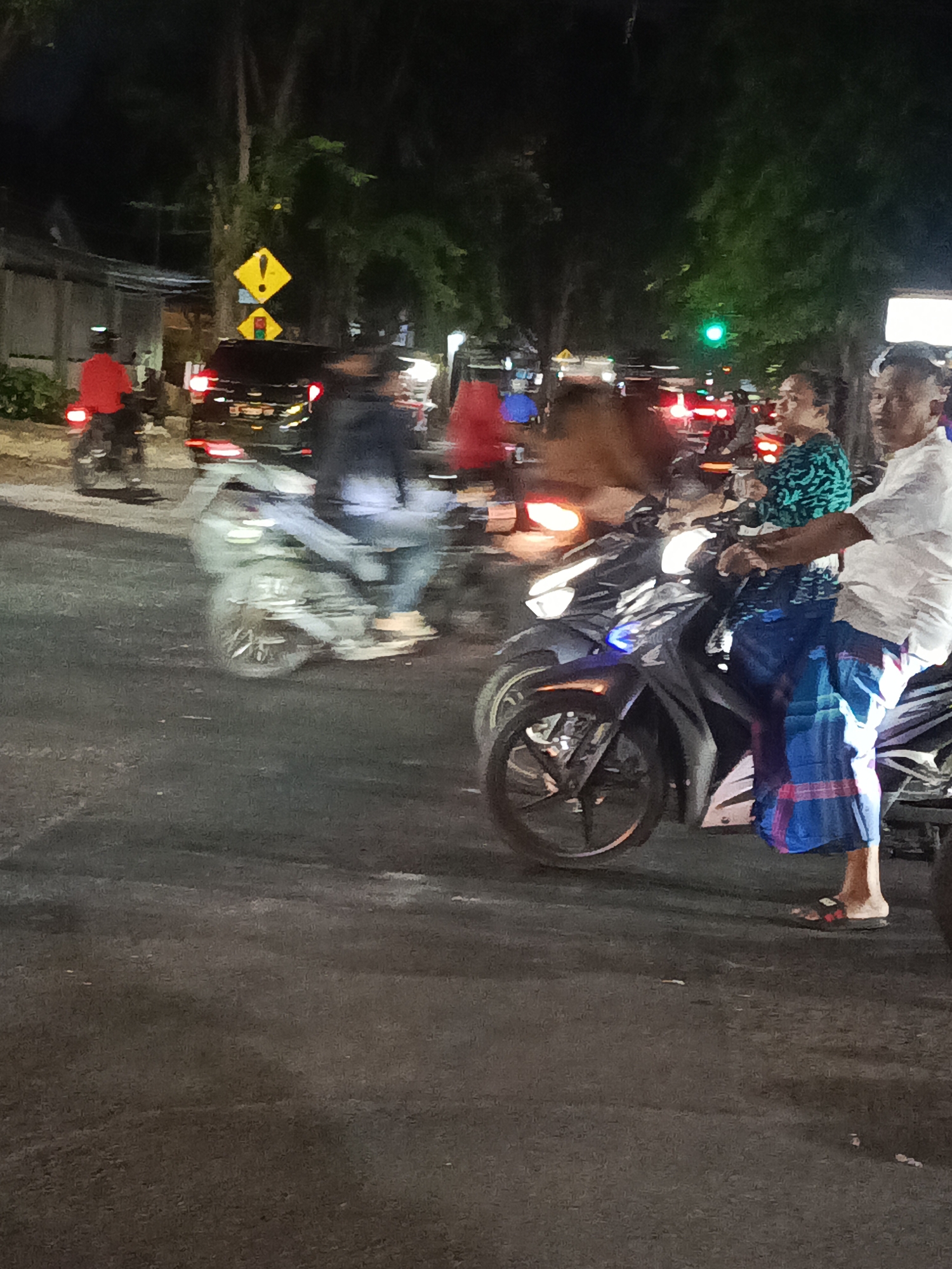 At night on a city street in Surabaya, Indonesia, two people are riding on a scooter. The person in front is wearing a green patterned shirt and a blue sarong. The person behind them is wearing a white shirt and a blue sarong with red trim. They are both facing forward and appear to be moving. The scooter is dark colored and has its headlights on. In the background, there are trees, streetlights, and other vehicles, suggesting a busy urban environment. A yellow diamond-shaped traffic sign with an exclamation mark is visible on the left side of the image, along with a smaller diamond-shaped sign below it that appears to be a traffic signal. The ground is asphalt, and there are some blurry streaks of light indicating movement.
