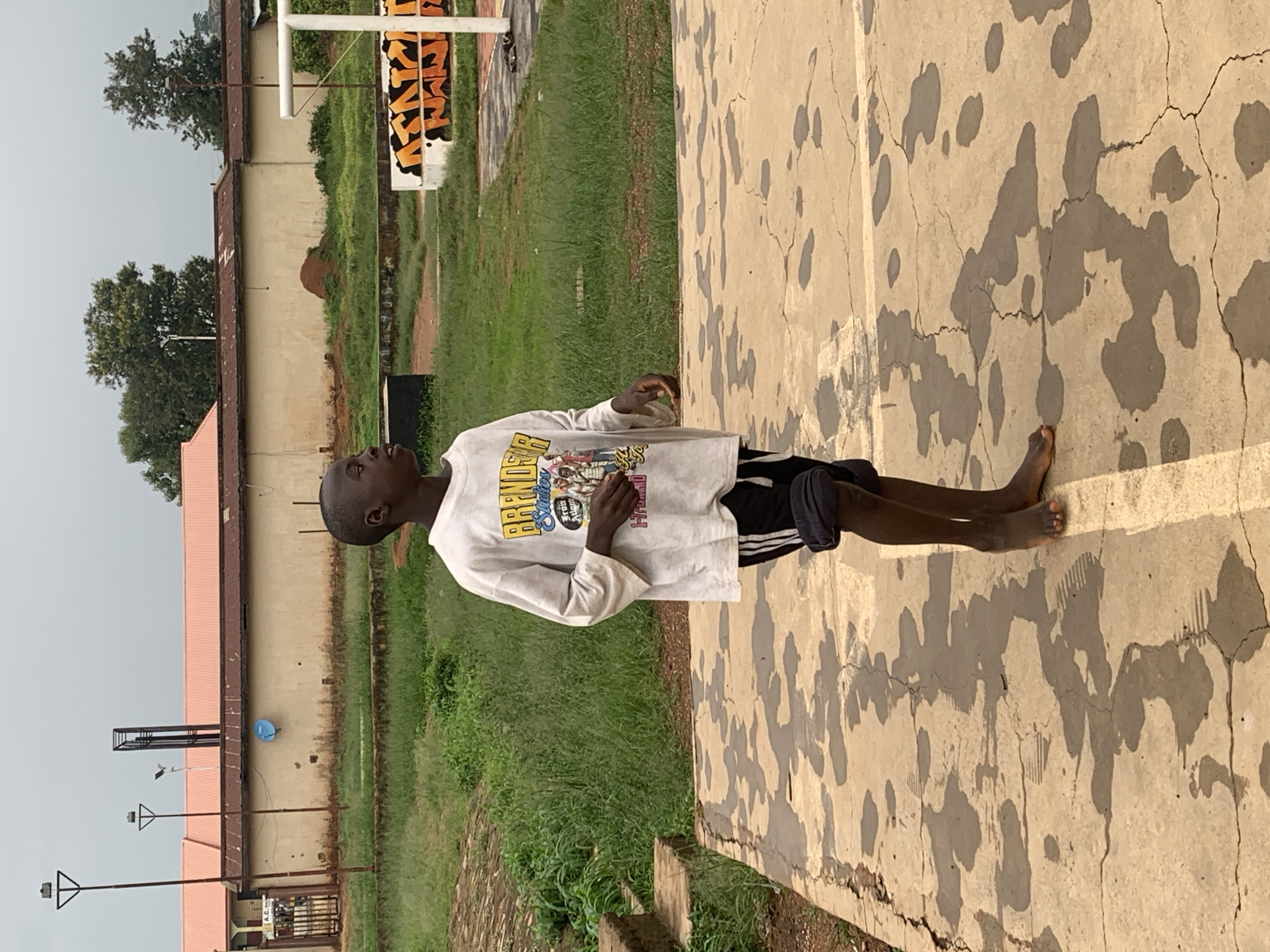 A young boy stands barefoot on a damp, cracked concrete surface, looking to his right. He is wearing a white t-shirt with a graphic design on the front and black shorts with white stripes. The t-shirt has text that appears to say "BRANDON Studios From Atlanta." The concrete surface is marked with dark, wet patches and some faint footprint impressions. In the background, there is overgrown green grass, a beige building with a red roof, and some trees. To the right of the frame, a white and orange structure, possibly a basketball hoop or goalpost, is visible. The overall setting suggests an outdoor recreational area in a somewhat unkempt urban environment. The scene is likely located in Jos, Nigeria, given the context provided.