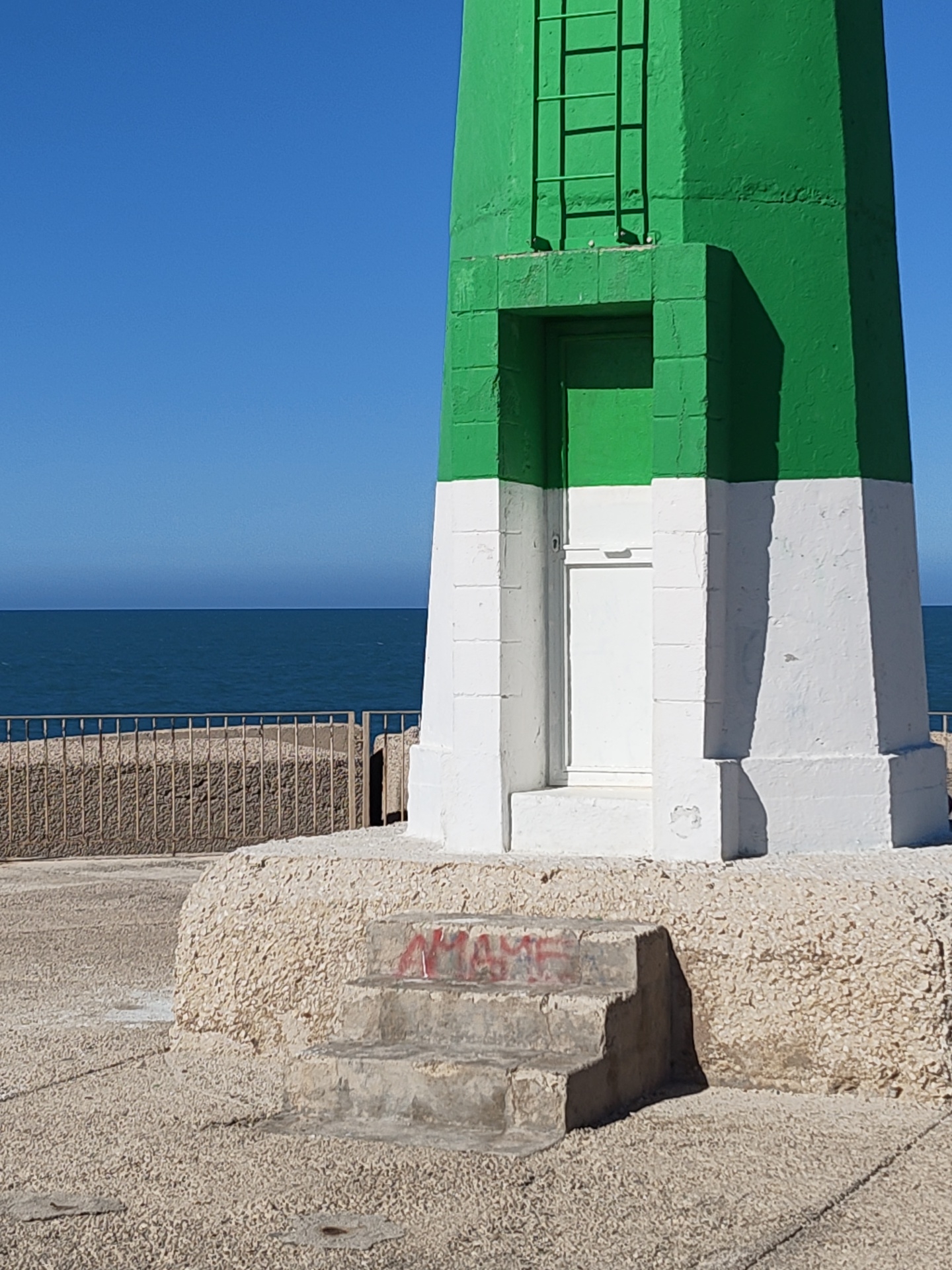 The image depicts a section of the green and white lighthouse at the end of Dénia's breakwater, Spain. The base of the lighthouse is comprised of a rough, light-colored stone, with a short flight of concrete steps leading up to a white door. There is a metal safety barrier visible in the background, and the sea and clear blue sky stretch out beyond. The time of day is likely daytime based on the clarity and sunlight present. No people are visible in the frame.