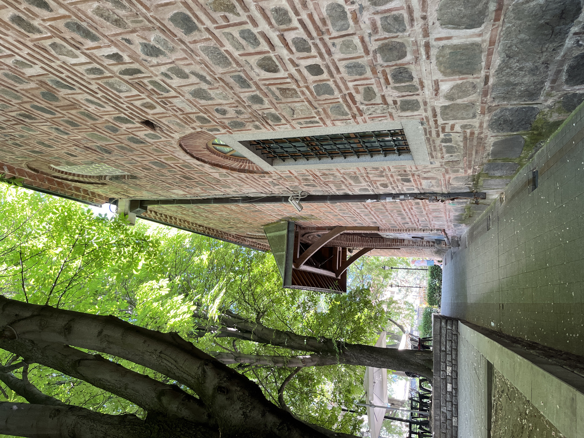 The image shows an exterior view of the Dzhumaya Mosque in Plovdiv, Bulgaria. The mosque's ceiling is brick with stone accents, featuring a rectangular barred window and a circular opening. An external wooden structure overhangs from the mosque, partially obscured by green foliage. A pathway runs alongside the mosque, with trees, greenery, and outdoor seating visible in the background. The overall scene is well-lit, suggesting daylight conditions.