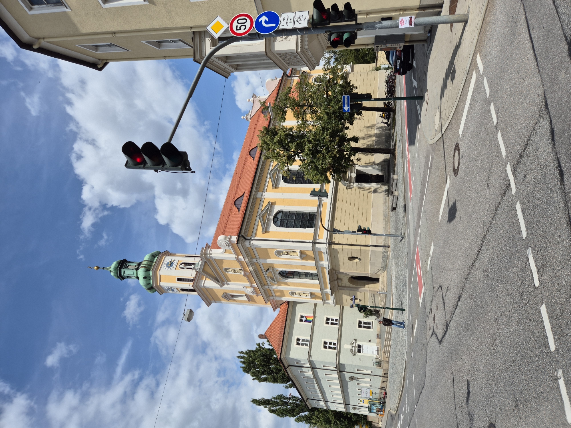 The image depicts a street view in Regensburg, Germany, featuring the St. Emmeram's Abbey, a prominent landmark. The abbey is characterized by its yellow facade, red tiled roof, and a distinctive green spire with a clock. In the foreground, a street intersection is visible with traffic lights, road markings, and a pedestrian crossing. A solitary person stands near the crossing. Adjacent to the abbey is a light blue building displaying a rainbow flag, suggesting a message of inclusivity. The sky is partly cloudy, indicating a bright day. The scene captures a typical urban environment with architectural elements, traffic infrastructure, and a touch of civic symbolism.