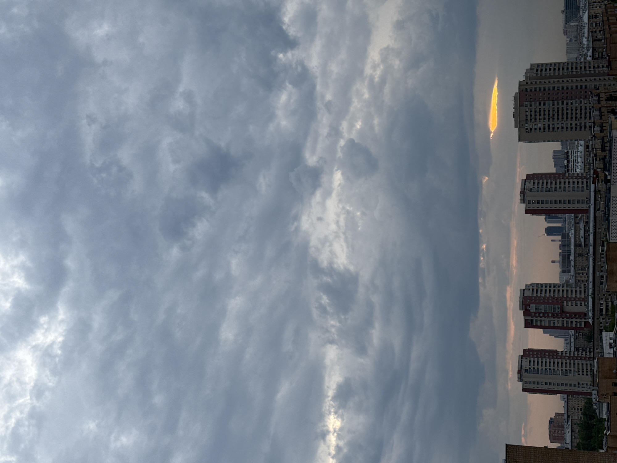 The image shows a view of Moscow, Russia, at twilight. The sky is filled with heavy, grey clouds, with a narrow vertical opening revealing a bright yellow sunset. Several tall residential buildings stand in the foreground, their facades showing red and white brick. The rooftops of lower buildings are visible. Green trees are interspersed between buildings.