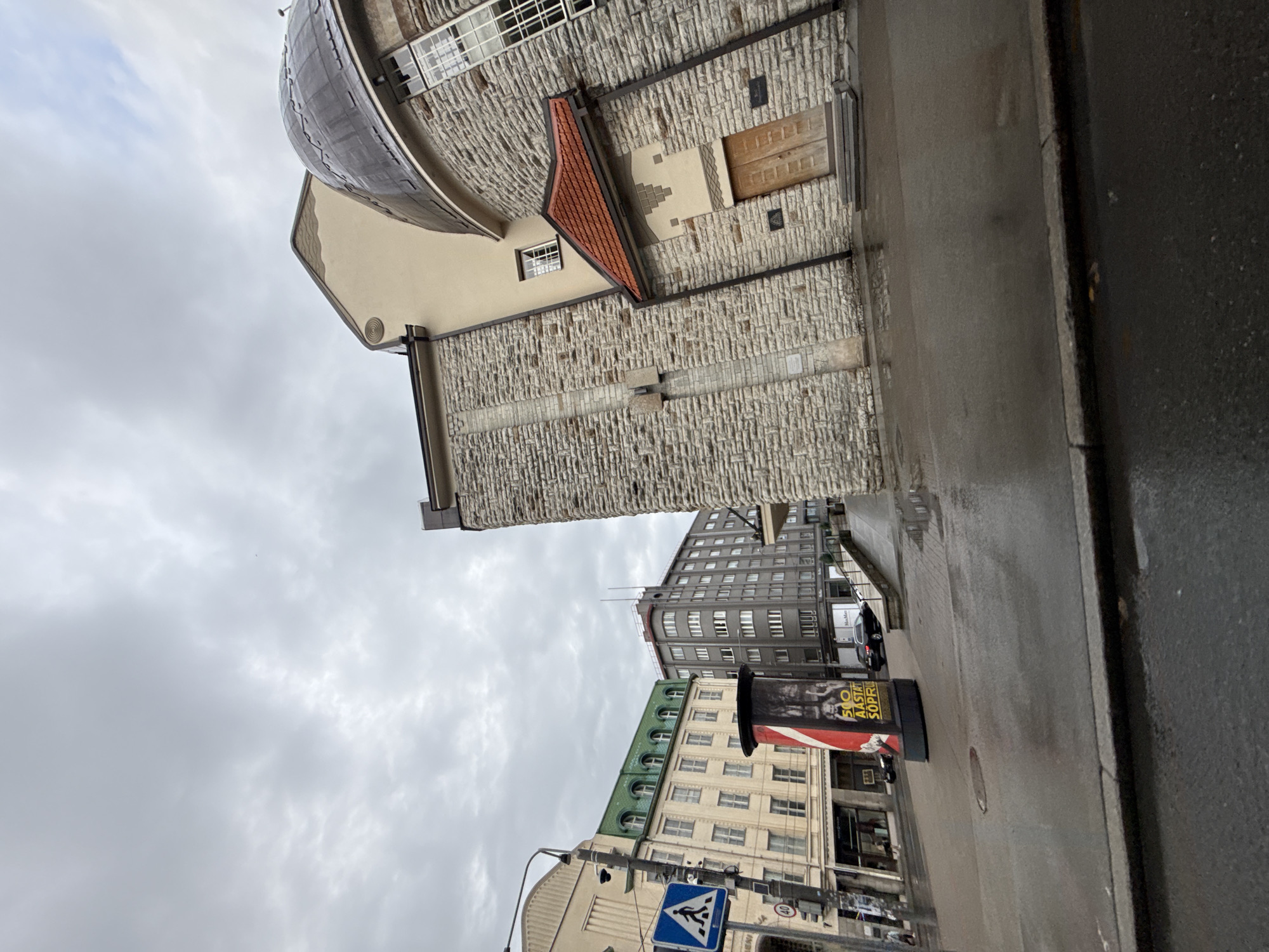 The image depicts a street scene in Tallinn, Estonia, on a cloudy day. On the right side of the frame, a building with a dark, domed roof and stone facade is prominent. The building has a red-tiled roof over an entrance with double wooden doors. To the left of this building, several other buildings are visible. One has a light-colored facade with a green, pitched roof and multiple windows. Further to the left, a taller, gray building with many windows is situated. In the foreground, a wet street reflects the overcast sky and buildings. A cylindrical advertising pillar with a red and white design stands on the sidewalk. Traffic signs, including a pedestrian crossing sign and a speed limit sign indicating 40, are visible on a lamppost. A black car is parked on the right side of the street, and a person is walking on the sidewalk on the far left. The overall atmosphere is somber, with the wet surfaces and cloudy sky suggesting recent or ongoing rain.