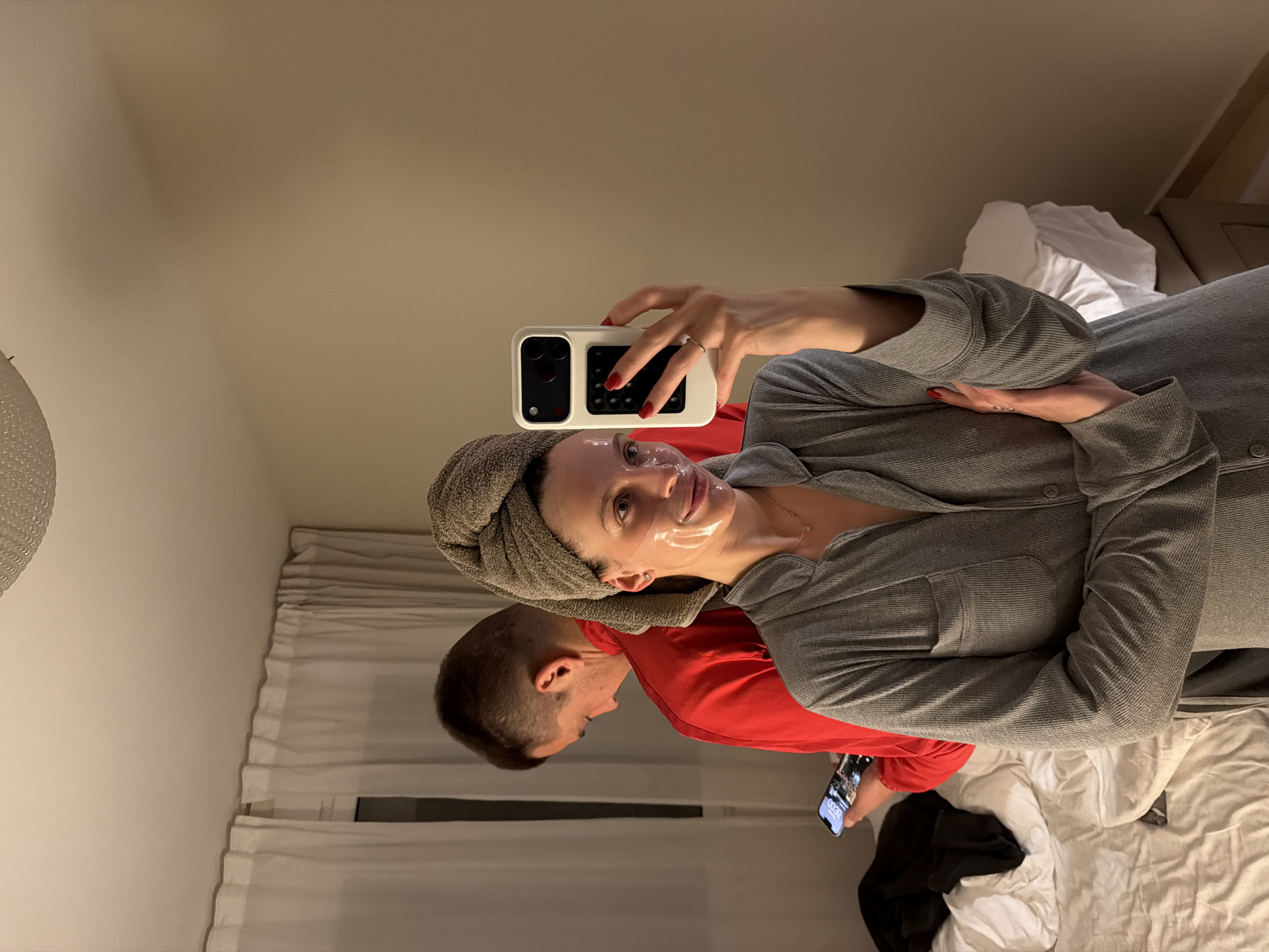 A woman with a towel wrapped around her head, wearing a clear face mask, is lying on a bed in a room. She is holding a white smartphone in her right hand, possibly taking a selfie. Her left hand rests on her stomach. She is wearing a grey button-down long-sleeved shirt. To her left and slightly behind her is a man wearing a red long-sleeved shirt, also lying on the bed. He is holding a black smartphone in his hand. The bed is covered in white linens, and there are dark clothes to the right of the bed. Behind the bed are curtains, and there is a beige wall with a bedside lamp. The setting suggests a hotel room or residence in Budva, Montenegro, although specific landmarks are not visible to confirm the exact location within the city. The scene conveys a relaxed atmosphere, possibly during a skincare routine or downtime.