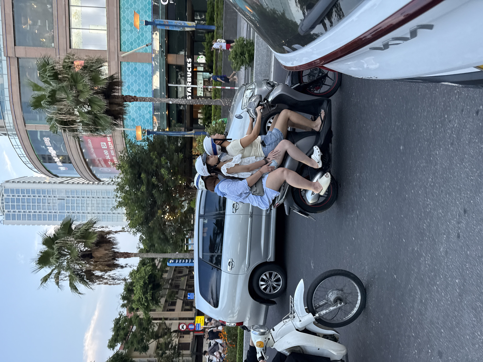 In Nha Trang Ward, Vietnam, a group of individuals navigates a city street on motorcycles. Two women are seen riding a scooter, with the driver wearing a white helmet and a denim skirt, and the passenger behind her also wearing a white helmet and a striped blue dress. To their left, another woman is on a separate white motorcycle. In the background, a silver van drives by, and a modern building with a large Starbucks sign is visible, hinting at a bustling urban environment. Palm trees line the street, adding a tropical element to the scene. The lighting suggests it is either early morning or late afternoon.