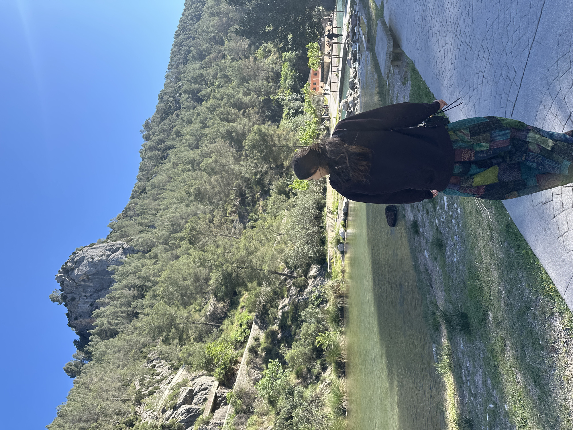 A full shot, slightly rotated to the left, shows a woman standing near a river and a cliff in Montanejos, Spain.
The woman, positioned in the right of the shot, is wearing a black sweater and a patterned skirt. She is facing away from the cliff and appears to be looking down at the river.
The river runs vertically through the center of the image, with green grass and foliage on its banks. A gray stone pathway is on the right.
On the left, a cliff covered in trees rises up to the sky. The sky is a clear blue. In the background, a bridge-like structure can be seen.