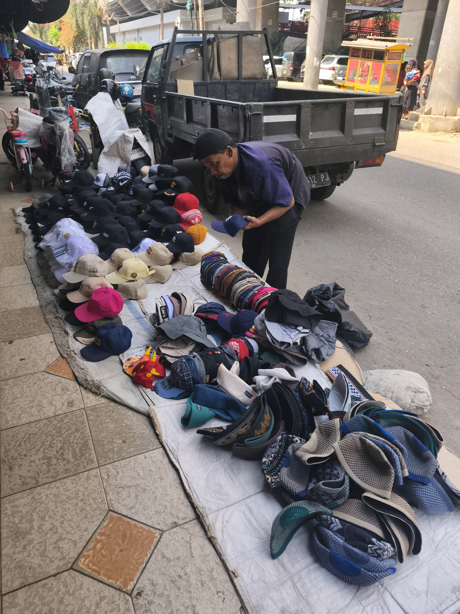 A street vendor in Kelurahan 16 Ilir, Kec. Ilir Timur I, Indonesia, is arranging a display of hats on the ground. The vendor, wearing a dark blue shirt and black pants, is bending over and holding a blue cap. The hats are laid out on a light-colored tarp on the sidewalk, bordering a paved street. The hats are diverse in color and style, with some featuring prominent logos. Behind the hats, several motorcycles and a black truck are parked. In the background, there is a yellow and red food stall, and pedestrian overpasses are visible. The overall scene depicts a bustling urban street market environment.
