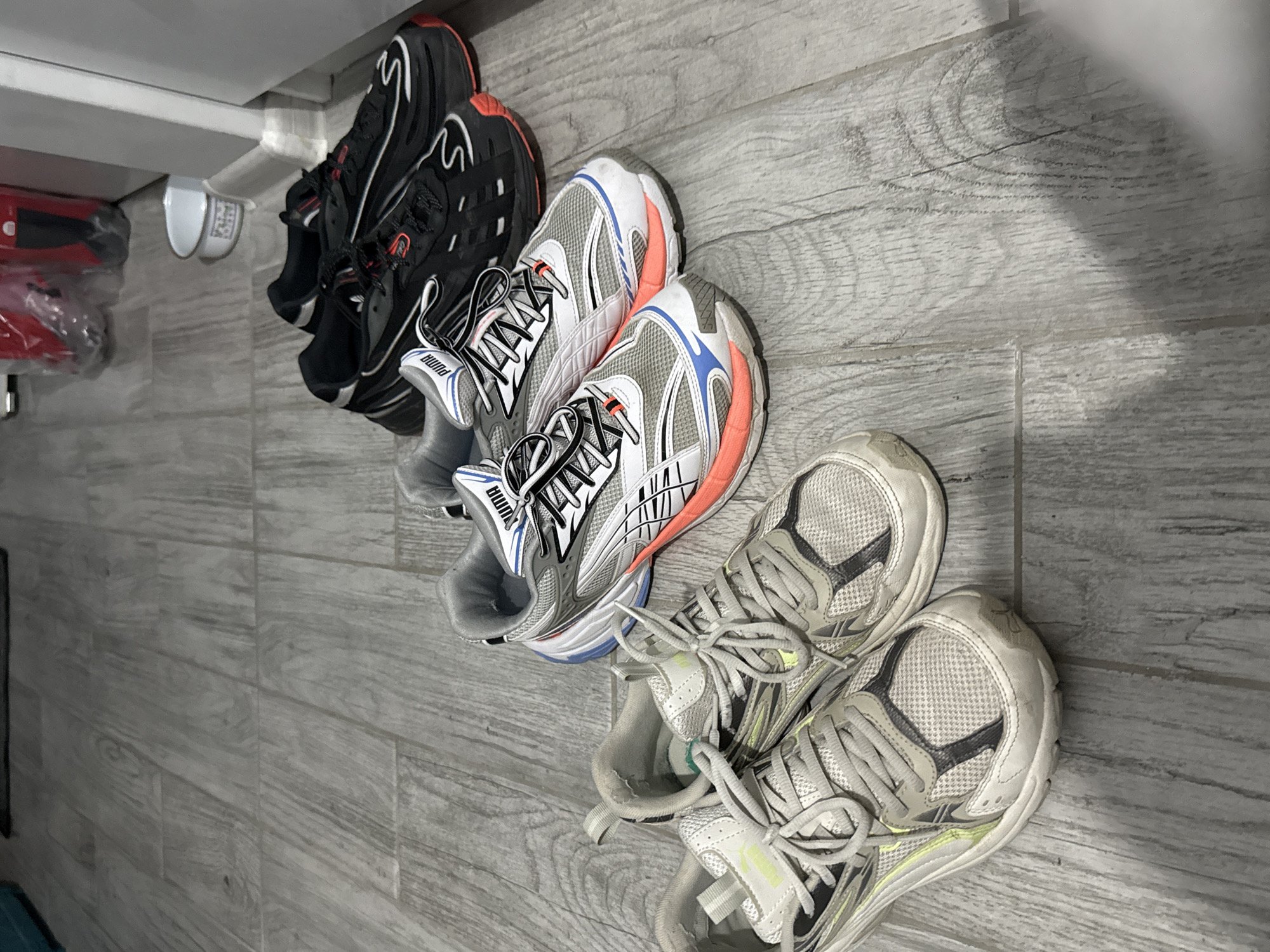 The image depicts three pairs of sneakers arranged on a light grey, wood-patterned floor. The sneakers are arranged in a row, with the toes pointed towards the right. From left to right, the first pair is black with red accents, the second pair is grey, white and orange, and the third pair is off-white with grey accents. The background shows a partial view of a white shelf. The location is likely indoors, in an apartment or house, and the city and country are not evident in the image.