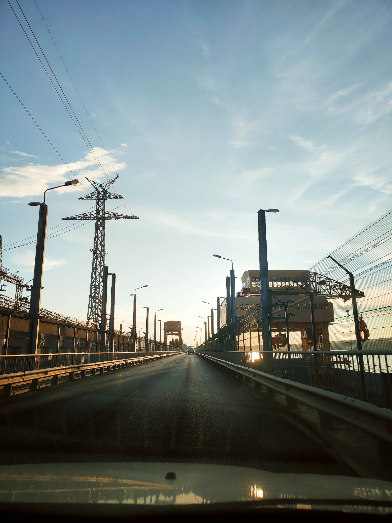 The image shows a view from inside a vehicle, looking forward onto a bridge or elevated roadway. The sky is clear with some wispy clouds, and the sun appears to be setting or rising, casting a warm glow. On either side of the roadway are industrial structures, including large metal pylons supporting power lines, streetlights, and a tall transmission tower. There is a fence made of wire mesh along the right side of the roadway. In the distance, the road continues and appears to lead towards a large, multi-story building or structure. The surface of the roadway is asphalt and appears wet in some areas, possibly from recent rain or mist. The reflection of the sky and structures can be seen on the dashboard of the vehicle in the foreground.