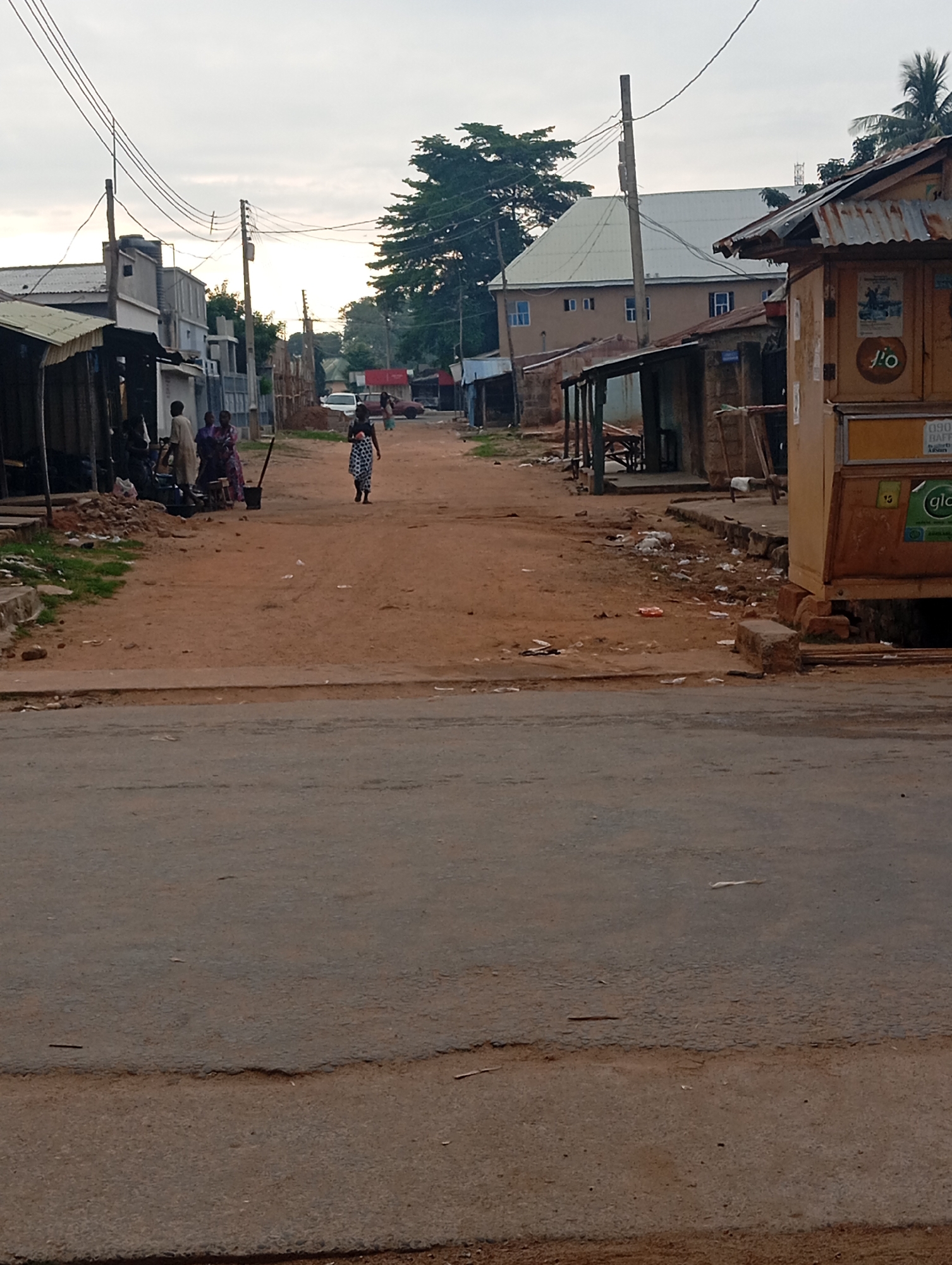 The image depicts a street scene in Jalingo, Nigeria, likely in a commercial or residential area. The foreground is dominated by an asphalt road with visible wear and tear. Beyond the road, a dirt path runs through the center of the scene, lined with structures on either side.
On the left side of the dirt path, several low-rise buildings with corrugated metal roofs are visible. Some appear to be market stalls or small shops, with open fronts and wares displayed. A group of people, including men and women in traditional attire, are gathered near these structures, some appearing to be engaged in conversation or activity.
On the right side of the dirt path, a more substantial, yellow-colored kiosk-like structure stands out. This structure has signage, including a "Glo" logo, suggesting it might be a telecommunications service point or a small business. Further down the street, more buildings and a large, light-colored building with a distinctive roofline are visible.
In the distance, a few vehicles, including a white car and what appears to be a red car, are parked or moving along the street. Several individuals are walking along the dirt path, including a woman in a patterned dress in the middle ground.
The environment appears somewhat untidy, with scattered debris and litter visible on the dirt path and along the sides of the road. Overhead, power lines crisscross the sky, supported by utility poles. The sky is overcast, suggesting either early morning or late afternoon. The overall impression is of a busy, everyday street scene in a Nigerian urban environment.