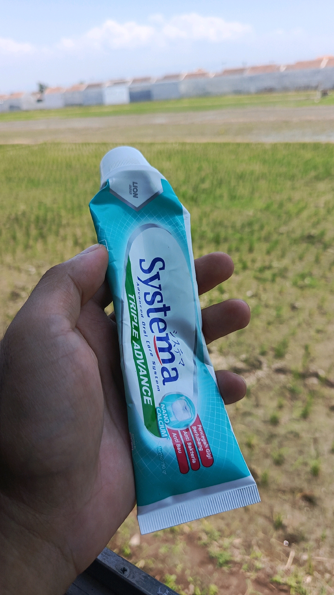 A hand holds a tube of Systema Triple Advance toothpaste in front of a blurred background of rice fields and houses under a bright, cloudy sky. The tube is primarily teal and white, with the brand name "Systema" prominently displayed in blue. Text on the tube indicates "Advanced Oral Care System" and mentions "Triple Advance," "Nano Calcium," and benefits like "Pencegah Gigi Berlubang" (Cavity Prevention), "Anti Bakteria" (Anti Bacteria), and "Anti Bau" (Anti-odor). The cap of the tube is white, and the brand "LION JAPAN" is visible near the top. The overall lighting suggests it is daytime.