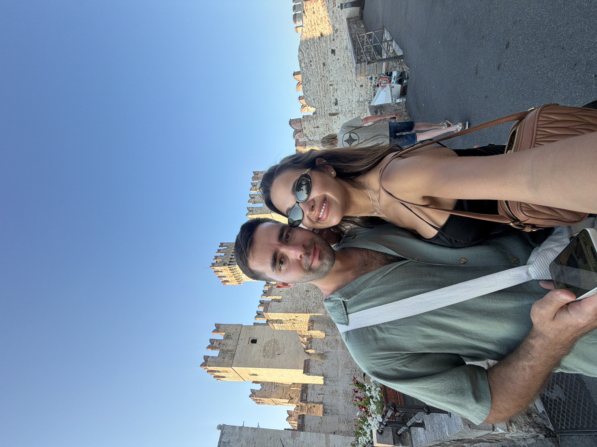 A couple takes a selfie in front of the Scaliger Castle in Sirmione, Italy. The man, in a green short-sleeved shirt, holds a smartphone. The woman, wearing a black top and sunglasses, smiles at the camera. Behind them, the ancient stone castle with its crenellated towers stands against a clear blue sky. A pedestrian walkway is visible to the right, with another person walking away from the camera. Flower pots line a ledge in the foreground.