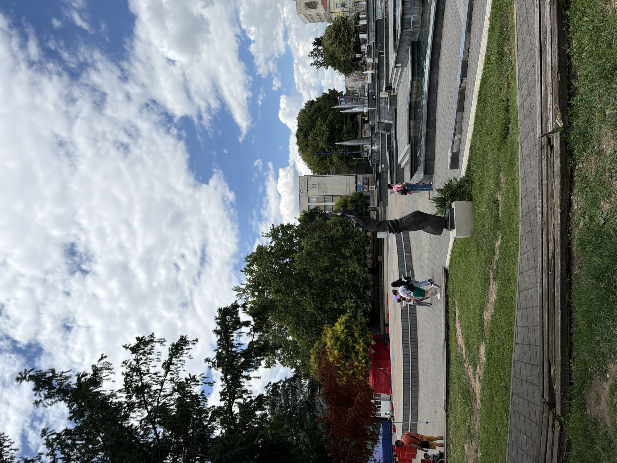The image depicts a public square with a fountain and a stage in Pazardzhik, Bulgaria. In the foreground, a grassy area with a wooden bench is visible. In the middle ground, a stage with a red tent behind it is set up. Several people are walking around the square. In the background, a complex fountain with multiple water jets is active. Buildings line the perimeter of the square. The sky is partly cloudy with a bright blue sky.