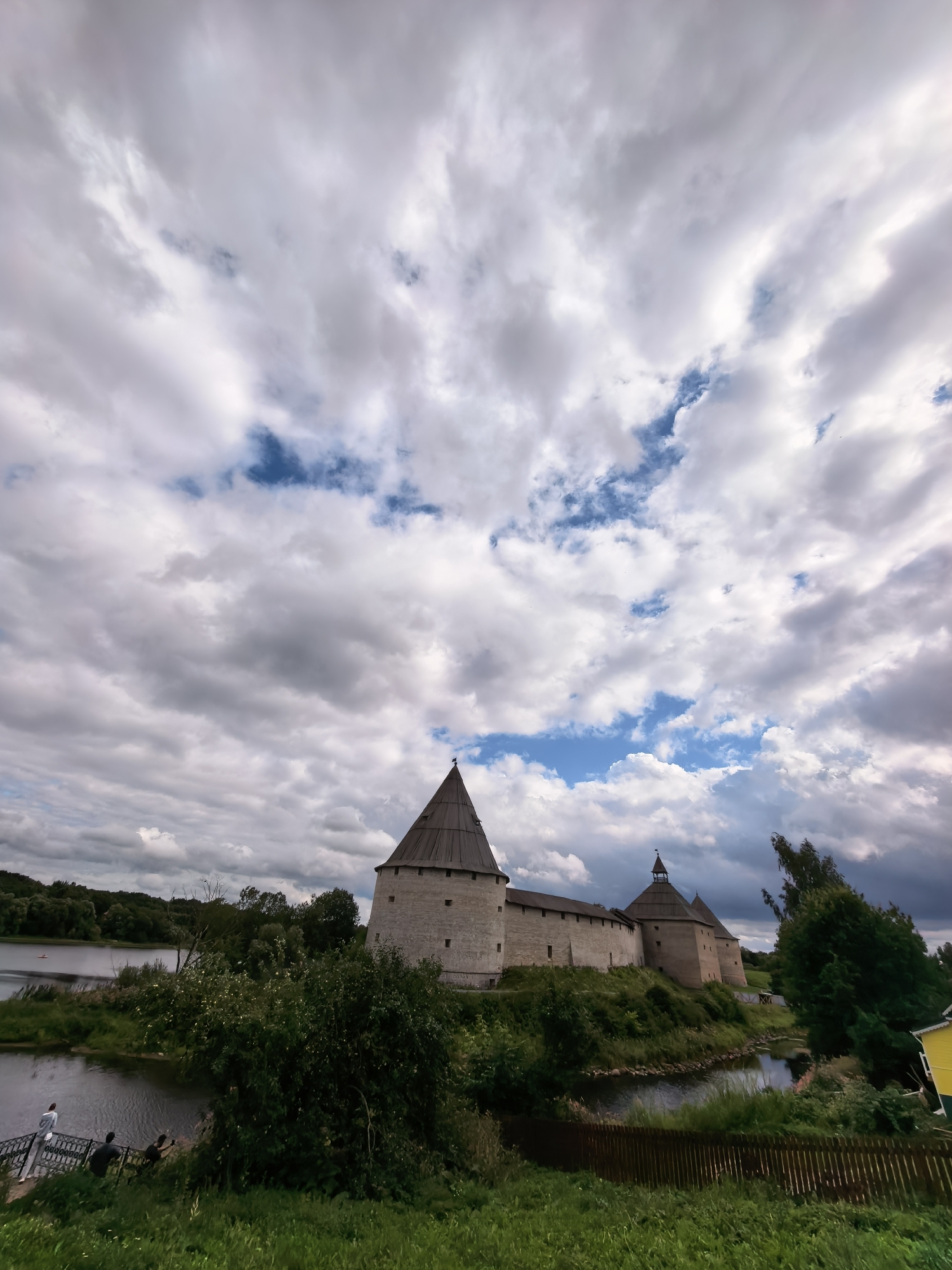 This is an eye-level, wide-angle shot of the Staraya Ladoga Fortress in Staraya Ladoga, Russia. The fortress, constructed of stone and featuring conical wooden roofs, is situated on a gentle slope overlooking a body of water. Two prominent towers are visible, one larger and closer to the foreground, and another slightly smaller one in the distance. The fortress walls stretch between them.
The water in the foreground reflects the dramatic, cloudy sky. On the left bank of the water, a metal railing with steps leads down to the water's edge. Three individuals are present near the railing: one person stands on the steps, and two others are seated on the ground beside them. A small boat is visible on the water in the distance.
The surrounding environment is lush and green, with abundant trees and foliage. Some trees are positioned in front of the fortress, partially obscuring it, while others line the far bank of the water. In the lower right corner of the frame, a section of a wooden fence runs along the edge of a grassy embankment. A small, bright yellow building with a dark roof is partially visible behind the trees on the right side of the image. The sky is filled with a mix of white and gray clouds, with patches of blue sky showing through.