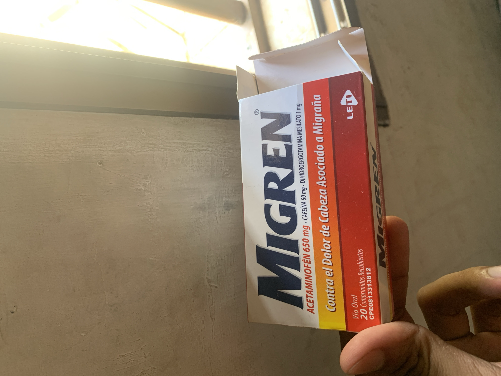 A hand holds an open box of Migren medication against a window in Santa Ana, Venezuela. The box is white with a red stripe; the box includes the drug's name, "Migren", and its use, "Contra el Dolor de Cabeza Asociado a Migraña." The window is to the upper left. The sun shines through the window and onto the side of the Migren box. The hand is in the lower right, obscuring part of the Migren box.