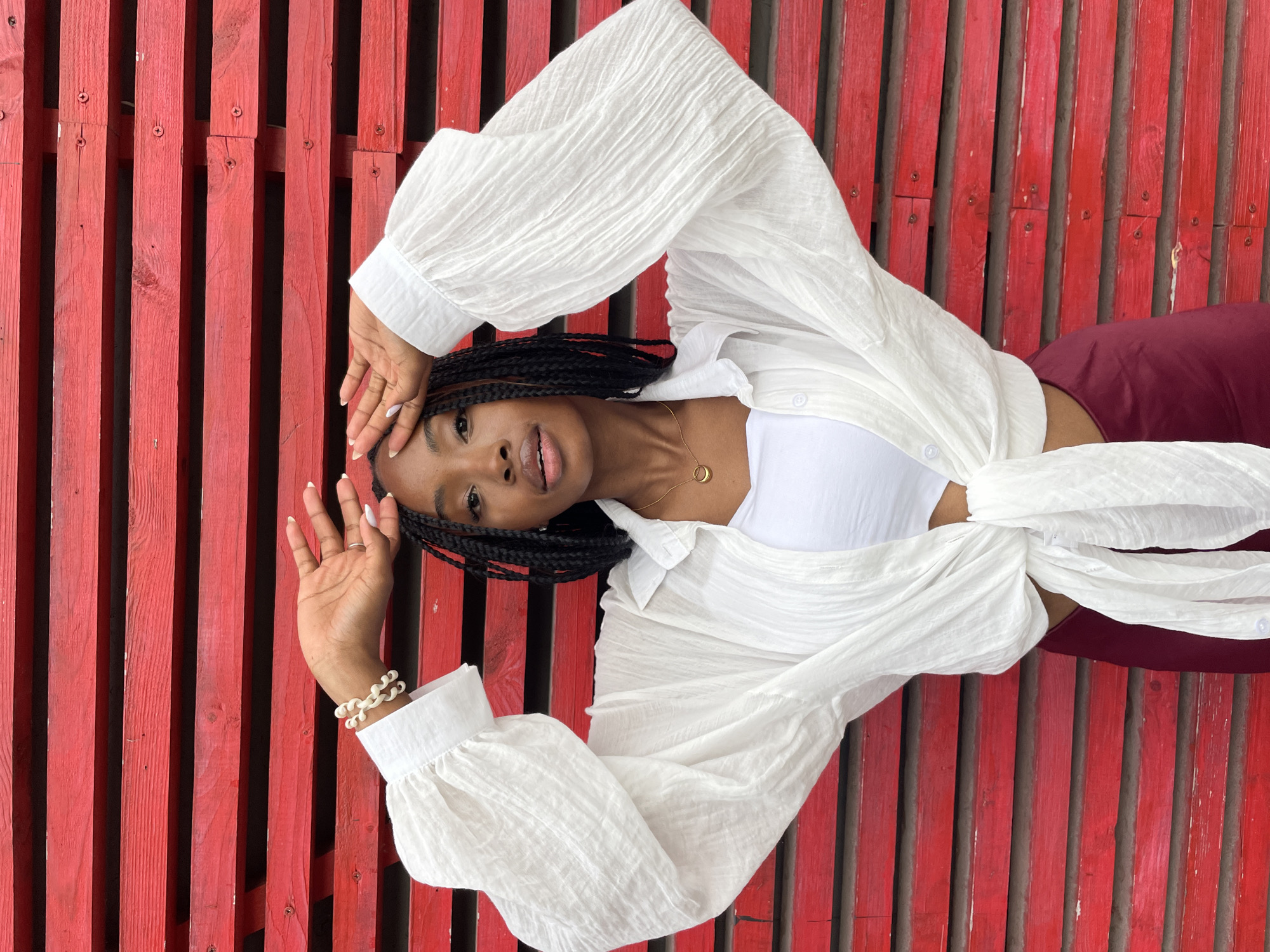 A young Black woman with braided hair stands in front of a bright red wooden wall. She is wearing a white, loose-fitting, long-sleeved shirt tied at the waist, over a white tank top. Her bottom garment is a maroon skirt. She has a gold necklace with a circular pendant. Her hands are raised to her forehead, and she is looking towards the camera. The background is a series of horizontal red wooden planks. The location is not specified to be in Kazan, Russia.
