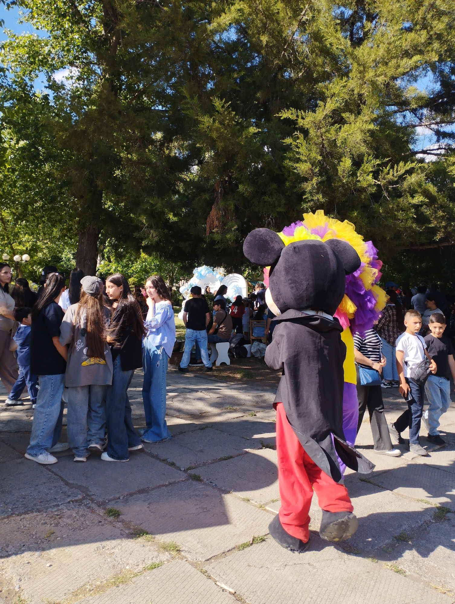 A daytime shot in Yeghegnadzor, Armenia, shows a street or plaza filled with people. In the foreground, a person in a Mickey Mouse costume stands with their back to the camera, wearing a black jacket, red pants, and oversized black shoes. A figure with a large, colorful, round headdress of yellow, purple, and white stands next to the Mickey Mouse character. A small group of young women in casual clothing stand to the left of the Mickey Mouse character. Behind them, other people are milling around; some stand, others sit. In the background, white balloons are visible. The area is paved with light-colored concrete slabs. The scene is set outdoors, with large trees providing shade.