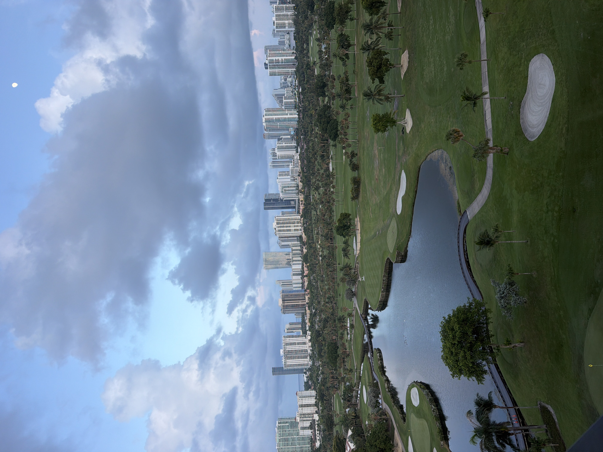 This aerial photograph captures a sprawling golf course in Aventura, Florida, USA, under a cloudy sky with a sliver of moon visible. The foreground features a meticulously maintained golf course with lush green fairways, strategically placed sand traps, and a winding body of water. A picturesque bridge crosses the water, connecting different parts of the course. Palm trees and other tropical vegetation are scattered throughout the landscape. In the background, a dense urban skyline is dominated by numerous high-rise residential buildings, indicative of Aventura's characteristic development. The scene appears tranquil, with no people or animals immediately visible on the golf course itself. The lighting suggests it might be early morning or late afternoon, given the soft light and the presence of the moon.