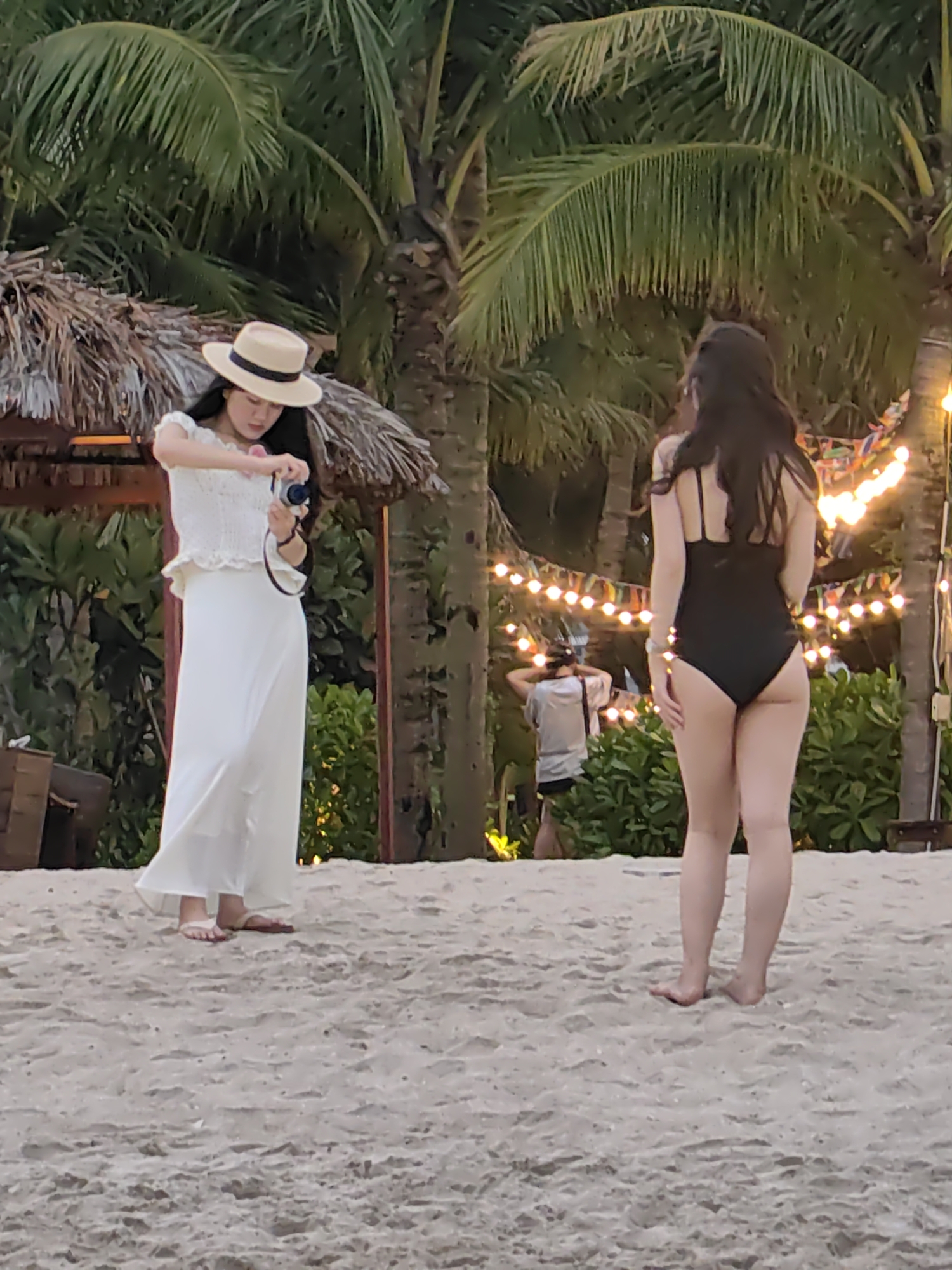 The image depicts a leisure scene on a sandy beach, likely at a resort in Bai Chay Ward, Vietnam, during the late afternoon or early evening.
In the foreground, two women are the primary subjects. On the left, a woman with long dark hair wears a light-colored wide-brimmed hat, a white ruffled off-the-shoulder top, and a long flowing white skirt. She is standing barefoot on the sand, holding a silver and black camera, and appears to be adjusting settings or reviewing an image. Her gaze is directed downwards at the camera. To her right, another woman, also with long dark hair, stands with her back to the viewer. She is wearing a black one-piece swimsuit and is barefoot on the sand, seemingly posing or waiting for a photograph to be taken.
In the midground, further behind the woman in the swimsuit, another person is partially visible, also holding what appears to be a camera or phone, suggesting multiple people are involved in photography or documenting the moment.
The background is rich with tropical elements, featuring several tall palm trees with lush green fronds. Behind the woman on the left, a thatched roof structure is visible, indicative of a beach bar or cabana. String lights with warm-toned bulbs are strung between palm trees, along with colorful triangular bunting, suggesting an festive or evening event atmosphere. Green foliage and bushes line the edge of the beach. The sand is light-colored and appears soft, with footprints visible. The overall mood is relaxed and recreational. There is no visible text in the image.