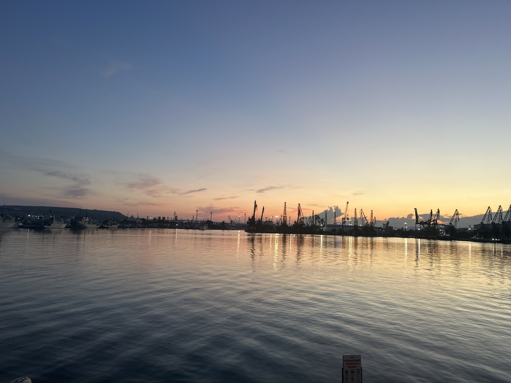 This image depicts a sunset over the Port of Varna, Bulgaria. The sky transitions from a deep blue at the top to shades of pink, orange, and yellow near the horizon, with wispy clouds scattered throughout. The foreground is dominated by calm water, reflecting the colors of the sky and the lights of the port. Along the shoreline, a silhouette of cranes, cargo ships, and buildings creates a stark contrast against the vibrant sky. A small, white sign with red lettering is visible in the lower right corner of the frame, indicating a warning or informational message. The overall scene is one of industrial activity set against a serene natural backdrop.