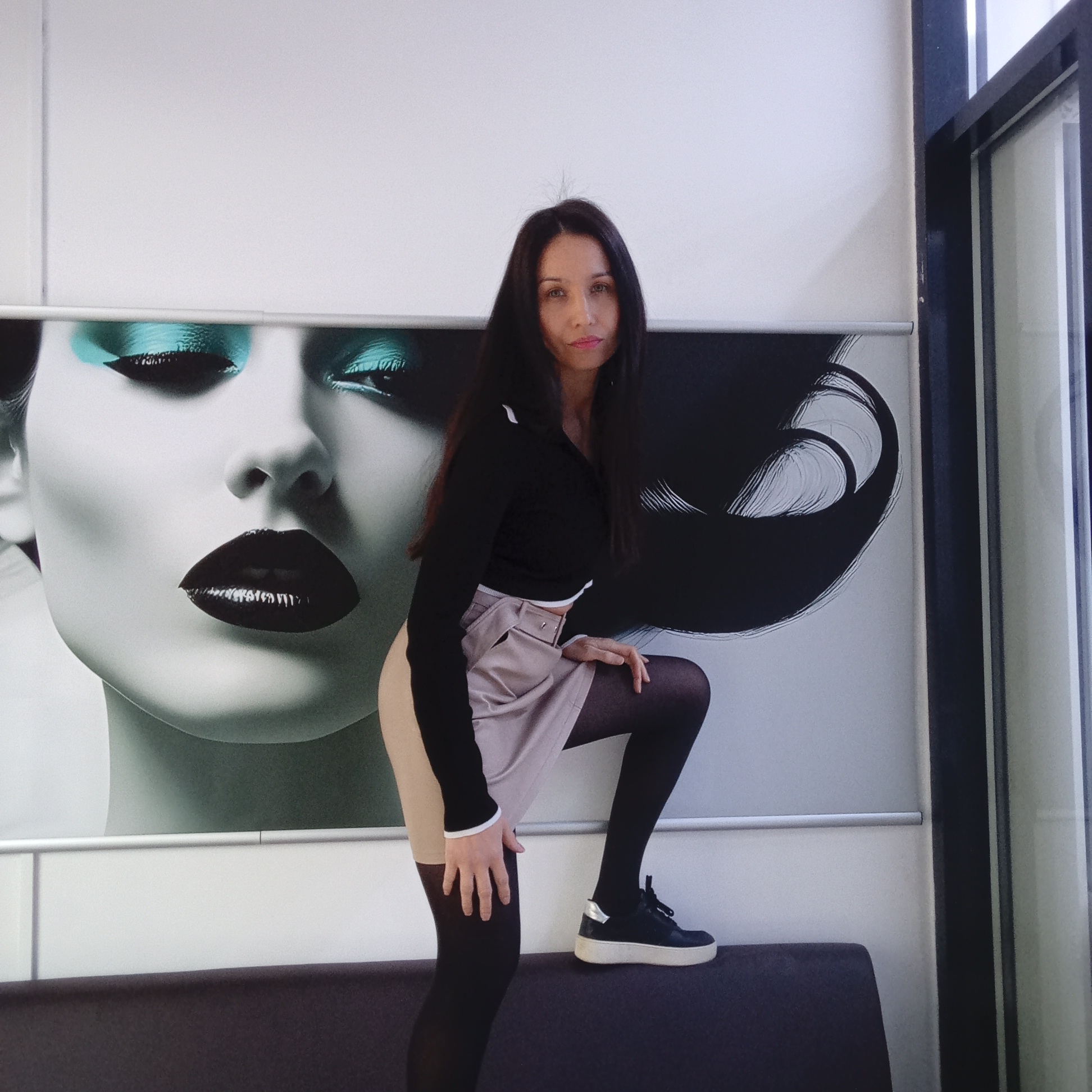 A woman poses in a vibrant indoor setting, likely in Izhevsk, Russia. She has long, dark hair and gazes directly at the camera with a neutral expression. She is dressed in a black long-sleeved top with white trim, a light beige or pale grey mini-skirt, dark opaque tights, and black platform sneakers with white soles and silver heel accents. Her pose is dynamic, with her left knee bent and foot resting on a dark upholstered bench or low partition, and her right leg slightly extended.
The background features a striking, large-scale mural or poster depicting a stylized close-up of a woman's face. The mural showcases vivid teal eyeshadow, dramatic black winged eyeliner, and glossy dark lipstick, set against a monochrome face. Abstract black and white lines flow around the facial image, suggesting artistic flair. Above the mural, the wall is plain white, and to the far right, a dark-framed window or glass partition is visible, hinting at an exterior view or another room. The lighting is bright and even, illuminating the scene clearly. The overall setting suggests a modern commercial space, possibly a beauty salon, studio, or a stylish lobby. There is no visible text in the image.