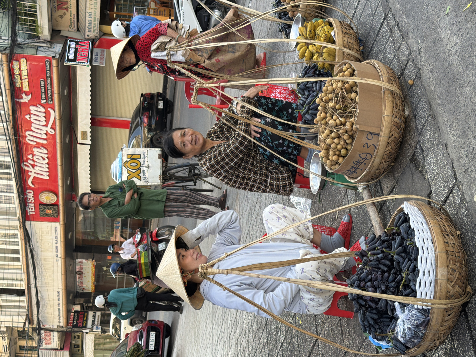 The image captures a street scene in Nha Trang, Vietnam, where two women are selling fruits from baskets. In the foreground, a woman wearing a conical hat and a light blue jacket sits on a red stool, her legs crossed, with a basket of dark, elongated grapes in front of her. To her right, another woman, dressed in a patterned shirt and dark pants, sits on a red stool, looking directly at the camera. She has a basket of what appear to be longans in front of her, and a basket of bananas to her right. Behind them, a man in a green shirt stands with his arms crossed, observing the scene. In the background, various shops line the street, indicated by signage. One prominent sign reads "thiên Ngân" in red lettering. Vehicles, including a red car and motorbikes, are visible, suggesting an active urban environment. The ground is paved with grey cobblestones. The overall atmosphere is that of a bustling marketplace.
