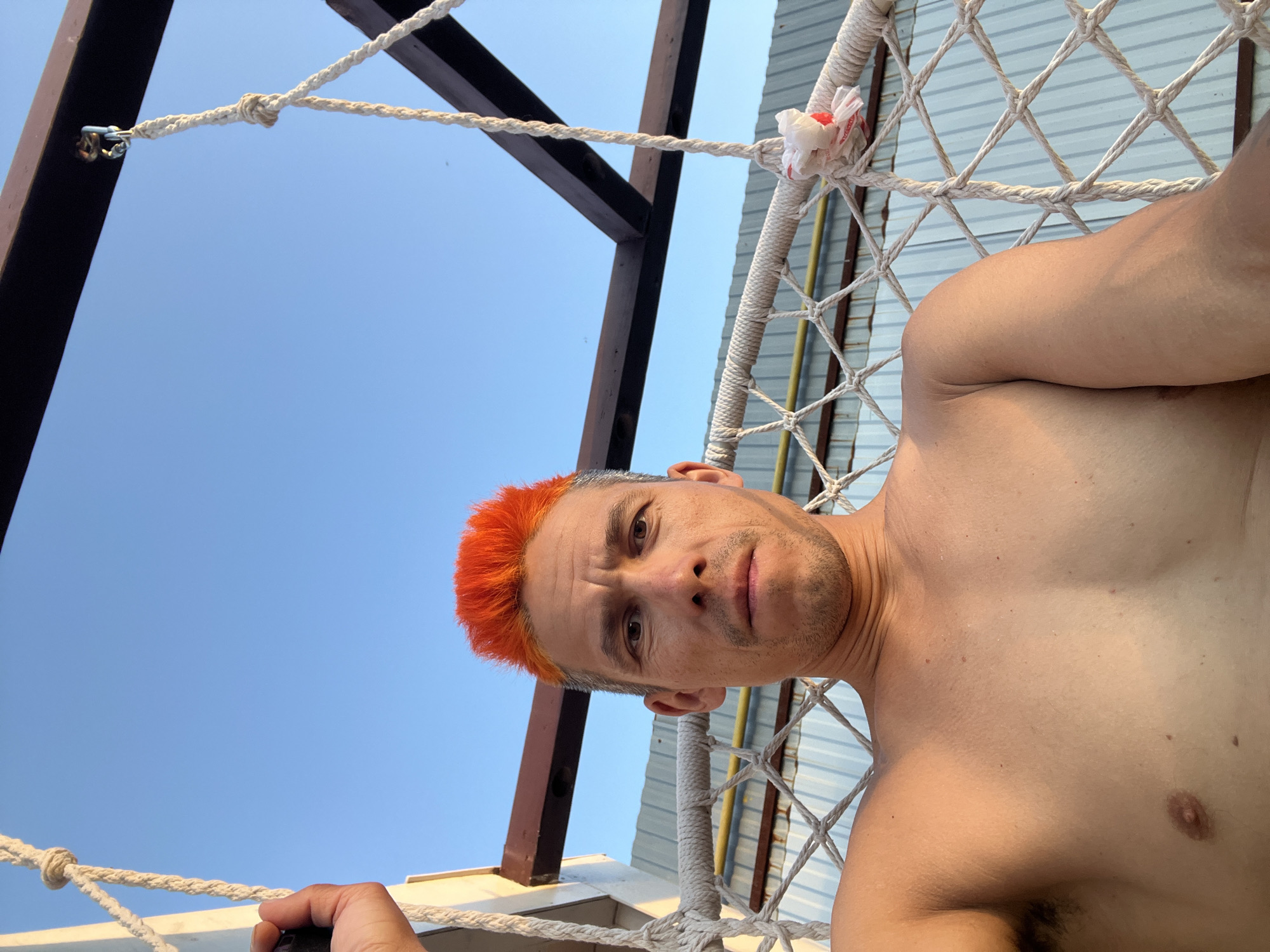 A low-angle, close-up shot depicts a shirtless man with orange hair lying in a hammock. The hammock is suspended from a dark wooden frame, with a white rope netting supporting the man. The background features a clear blue sky and a light-colored, corrugated metal wall. The man is looking directly at the camera. It is impossible to confirm that the picture was taken in Chaikovsky, Russia, without additional information.