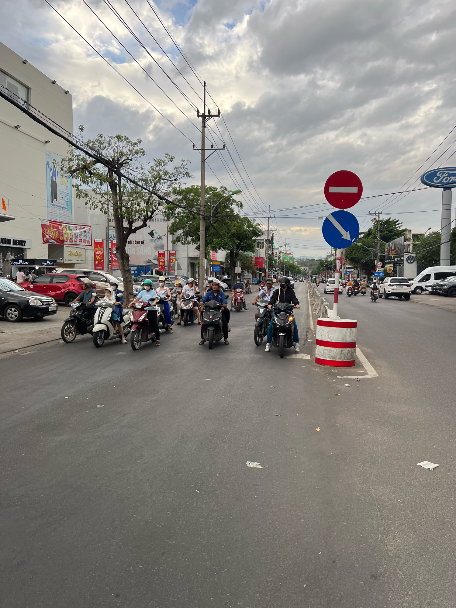 The image depicts a street scene in Nha Trang, Vietnam, likely during daylight hours with overcast skies. A multi-lane road is visible, with a significant number of motorcycles and scooters navigating through traffic. Several individuals are riding these vehicles, many wearing helmets and some with face masks. Cars, including a red sedan and a black sedan, are also present on the road.
On the left side of the road, a modern building with a large white facade is visible, featuring signage and posters. Trees line the street, and various utility poles and overhead wires crisscross the sky. On the right side of the road, a white Ford dealership sign is noticeable, along with other businesses. Traffic signs are visible, including a "no entry" sign (red circle with a white horizontal bar) and a directional sign indicating a downward arrow in a blue circle. A concrete barrier with red and white stripes is positioned near the road. The road surface appears to be asphalt and shows some signs of wear and debris. The overall impression is a busy urban environment with common modes of transportation and commercial establishments.