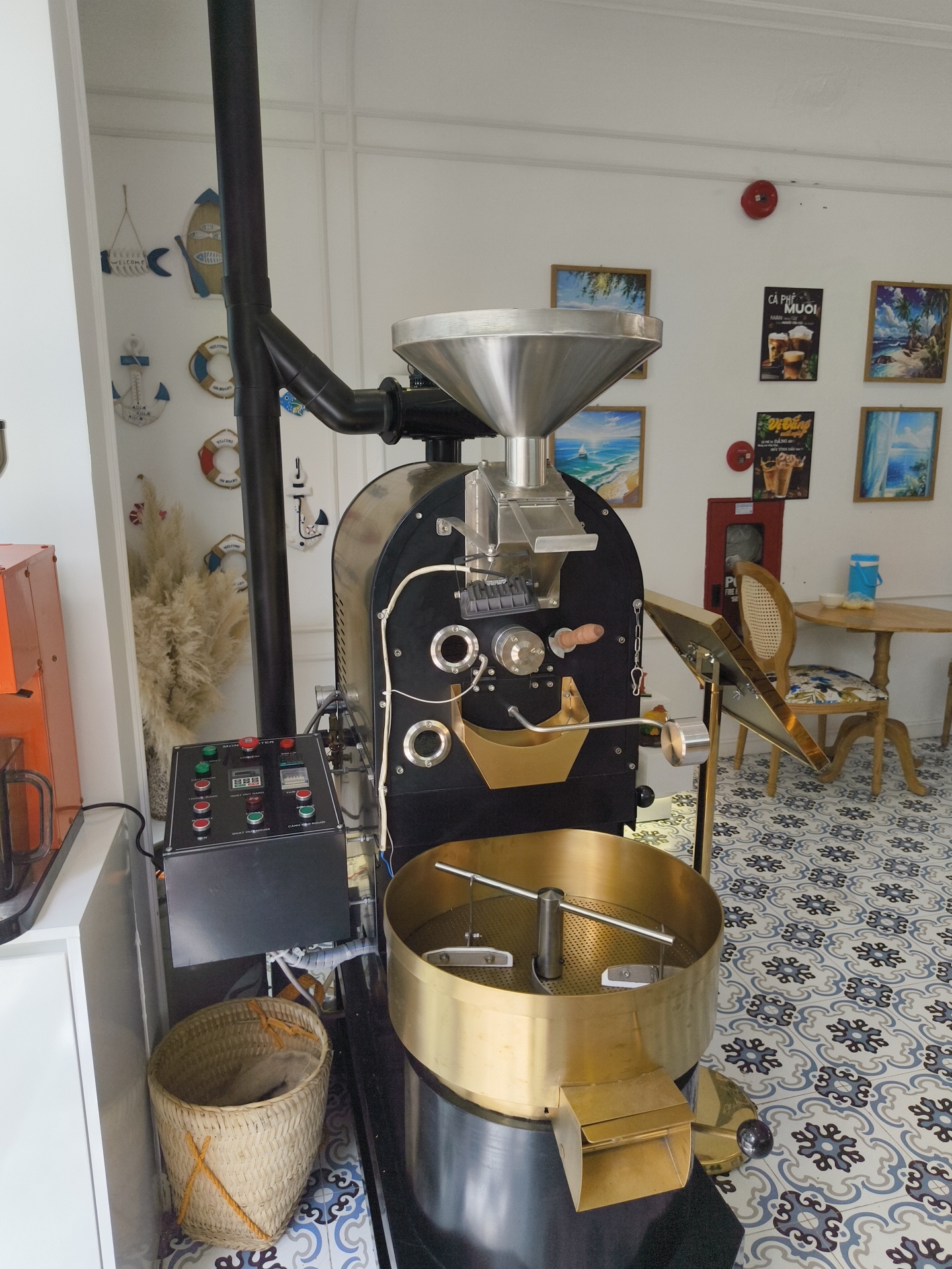 The image shows a coffee roasting machine in what appears to be a cafe or shop, likely located in Phú Quốc, Vietnam. The machine is black and stainless steel with a large silver hopper on top. To the left of the roaster, there is an orange piece of equipment, possibly another coffee-related machine. A control panel with buttons and digital displays is situated below the roaster. A woven basket filled with coffee beans sits on the floor beside the control panel. The background features a white wall decorated with framed pictures of beach scenes and nautical-themed ornaments such as life preservers and anchors. There is also a glimpse of a wooden table and chairs, suggesting a seating area for customers. The floor is tiled with a decorative pattern in shades of blue and grey. The overall impression is a well-lit, tidy establishment with a focus on coffee.
