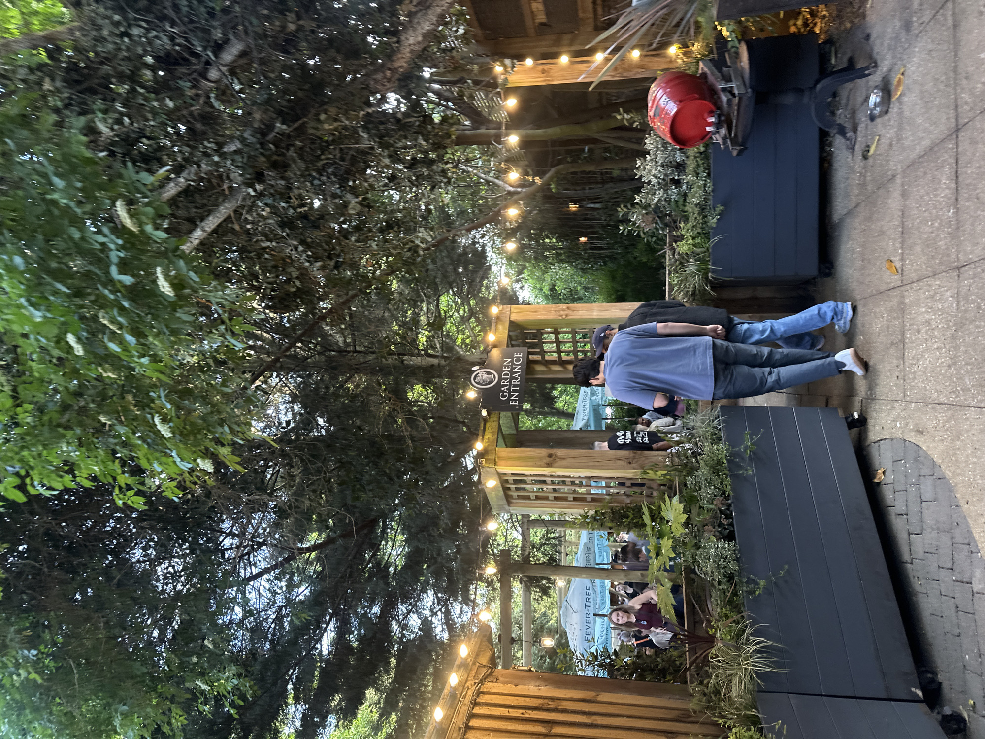 Here is a detailed description of the image:
**Setting and Environment:** The image shows an outdoor area, possibly a garden or entrance to a venue called "Garden Entrance." It has a rustic, slightly whimsical feel with wooden structures, overhead string lights, and abundant greenery. The scene appears to be at dusk or twilight, based on the lighting. The brand "Fever-Tree" is visible on signs within the space.
**People:** Several people are visible in the image. A few are standing at the "Garden Entrance" sign, looking into the venue. Other people can be seen sitting at tables inside the garden area, likely patrons of the venue.
**Objects:** Some primary objects visible include:
* Wooden structures forming an entrance or pergola.
* String lights illuminating the area.
* Planters with greenery.
* A red container or object mounted on a platform, placed near the entrance.
* Signs with the venue name ("Garden Entrance").
* Signs indicating "Fever-Tree."
**Actions:** People are primarily either entering the garden, standing near the entrance, or seated inside, suggesting social interactions and enjoyment of the venue.
**Location Clues:**
While there is no explicit indication of the location being London, United Kingdom, the signs for "Fever-Tree" and the architectural style of the garden elements are more commonly found in the United Kingdom.