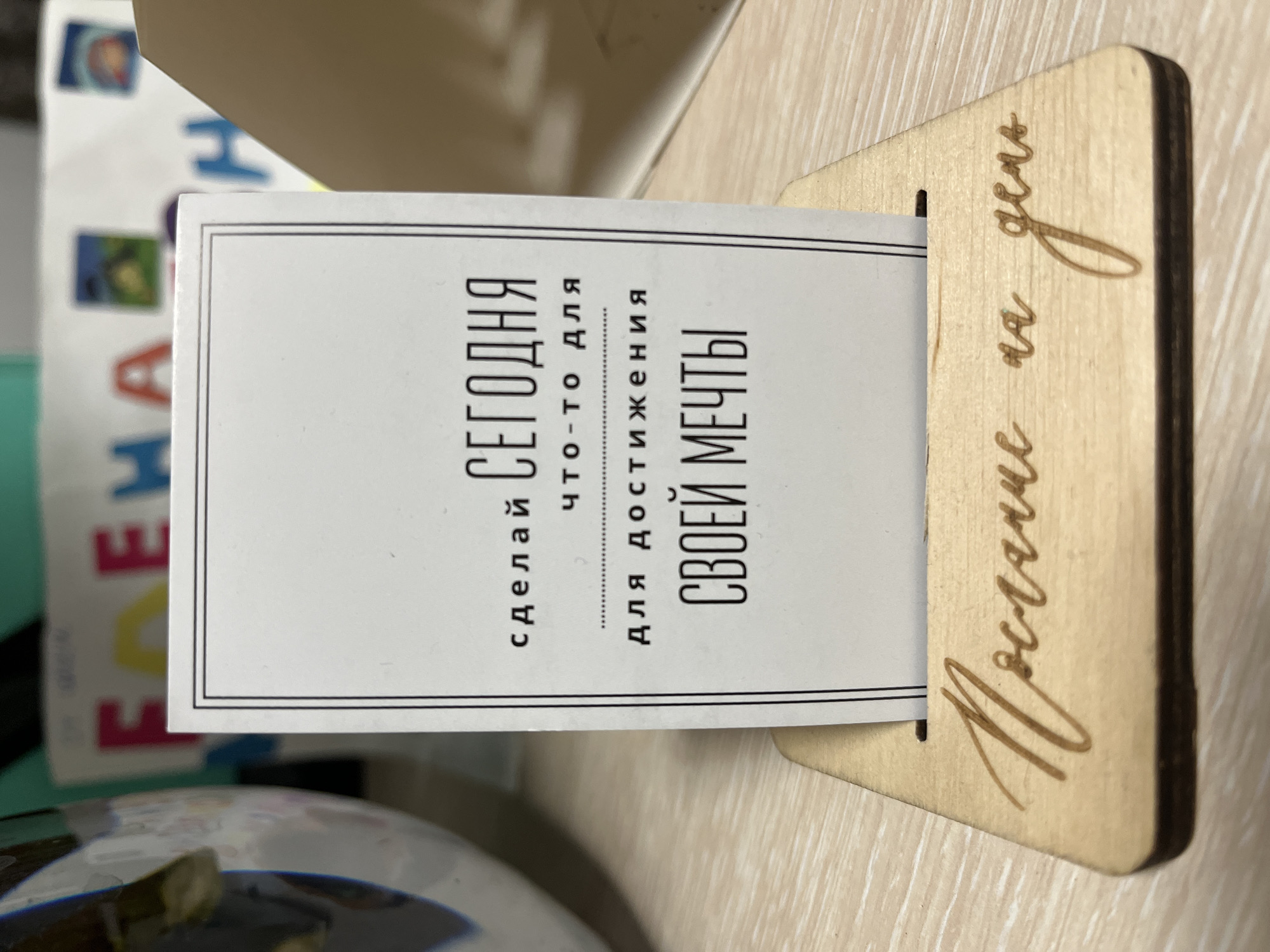The image shows a wooden stand with a card inserted into it. The card is white with black text. The text on the card reads: "СДЕЛАЙ СЕГОДНЯ ЧТО-ТО ДЛЯ ДОСТИЖЕНИЯ СВОЕЙ МЕЧТЫ" which translates to "DO SOMETHING TODAY TO ACHIEVE YOUR DREAM." Below the card, also on the wooden stand, is a handwritten inscription that reads "Постулат на день" which translates to "Motto for the day." The background of the image is out of focus but appears to be a table with some other items on it, including what looks like a colorful banner with letters. No specific location in Petropavlovsk-Kamchatsky, Russia can be determined from the image. There are no people or interactions visible in the image.
