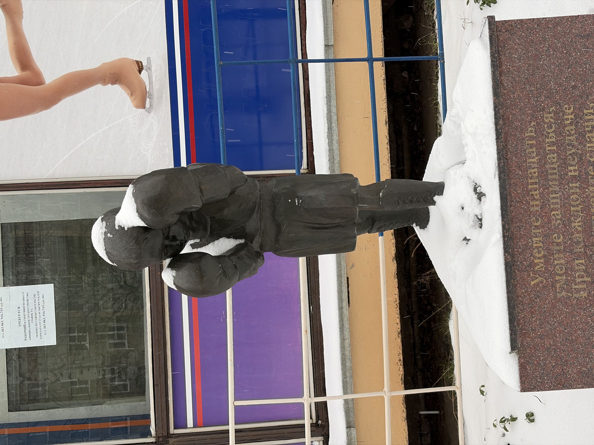 The image shows a statue of a figure skater with their head bowed in front of a granite monument partially covered in snow. The monument has text written in Russian, which is partially obscured. Behind the statue, there is a window with a poster on it advertising job opportunities in a sports school. To the left of the window, there is a depiction of a pair of ice skates on ice. Behind the statue and monument, there is a metal fence and some greenery in the background. The location is believed to be in Izhevsk, Russia, based on the text on the monument, which mentions Izhevsk.