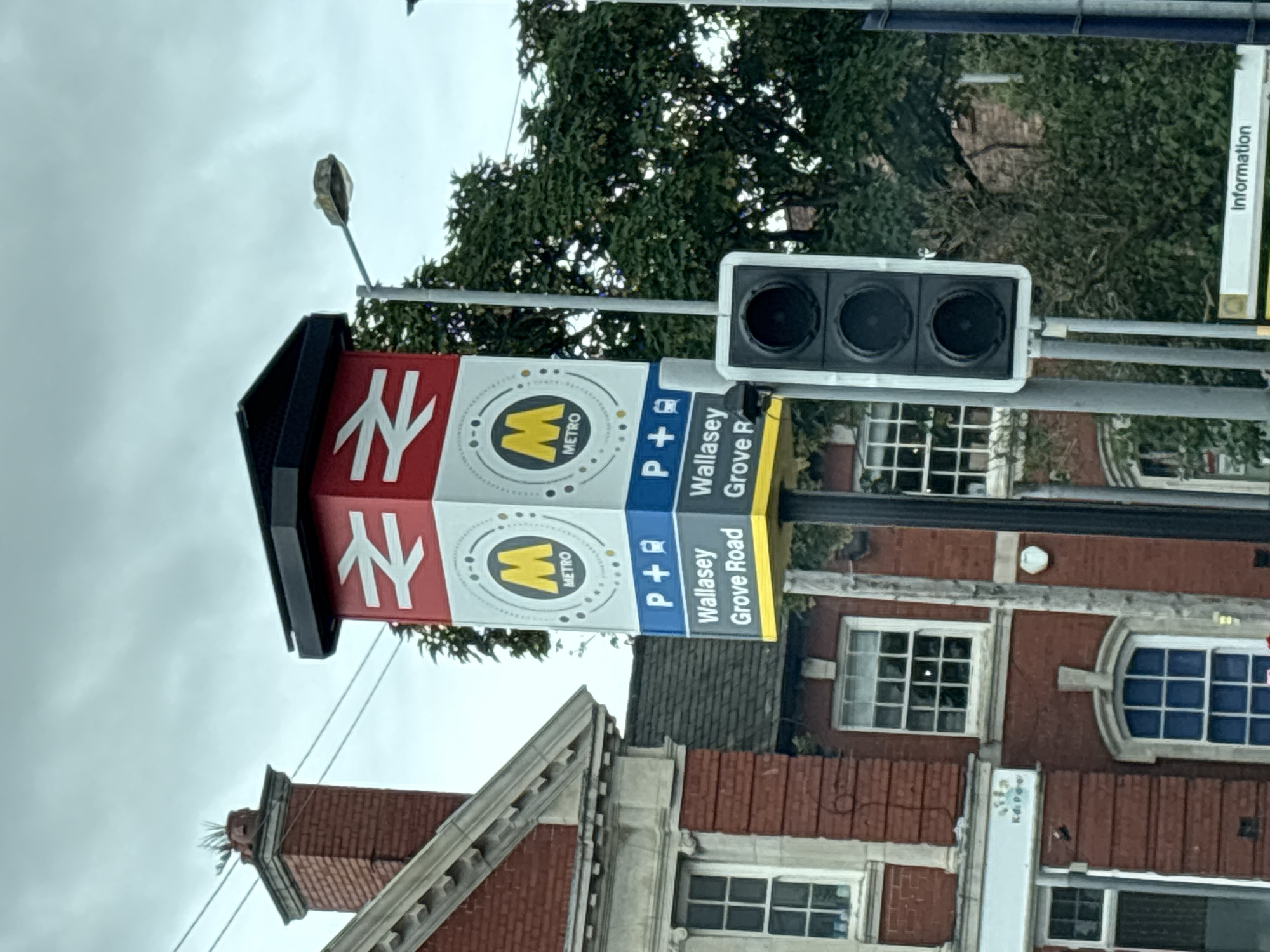 The image shows a signpost for Wallasey Grove Road station, a Merseyrail station in Wallasey, United Kingdom. The sign is red and white and features the Merseyrail logo. There is also a traffic light and a street lamp visible in the image. The station building is made of red brick and has several windows. Trees are visible in the background. The sky is overcast.