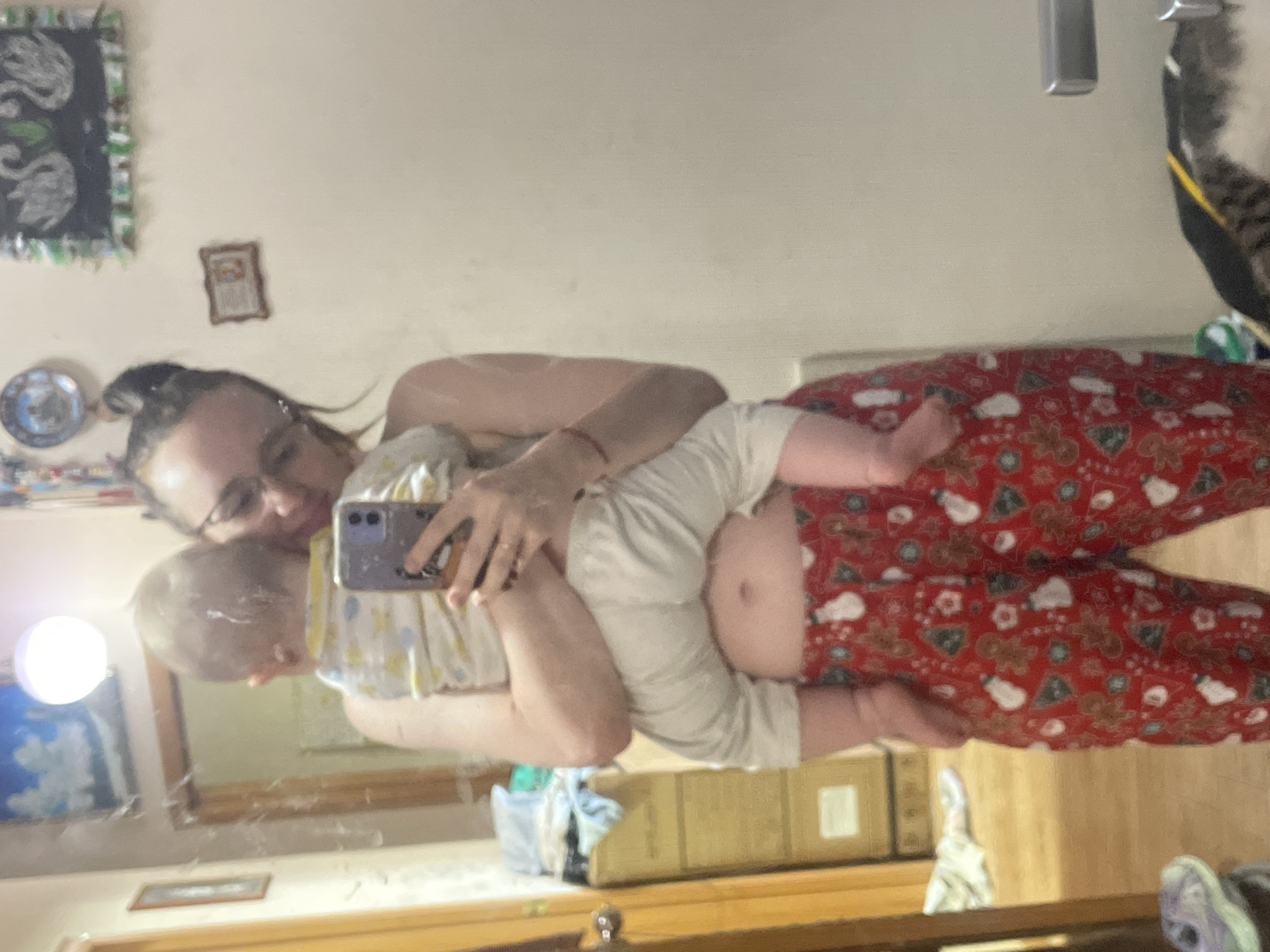 A woman is holding a baby and taking a selfie in a mirror. The woman is wearing glasses and has her hair up. The baby is wearing light-colored clothing. The woman is wearing red patterned pajamas. The environment appears to be a bedroom. The location is Unknown, Unknown.