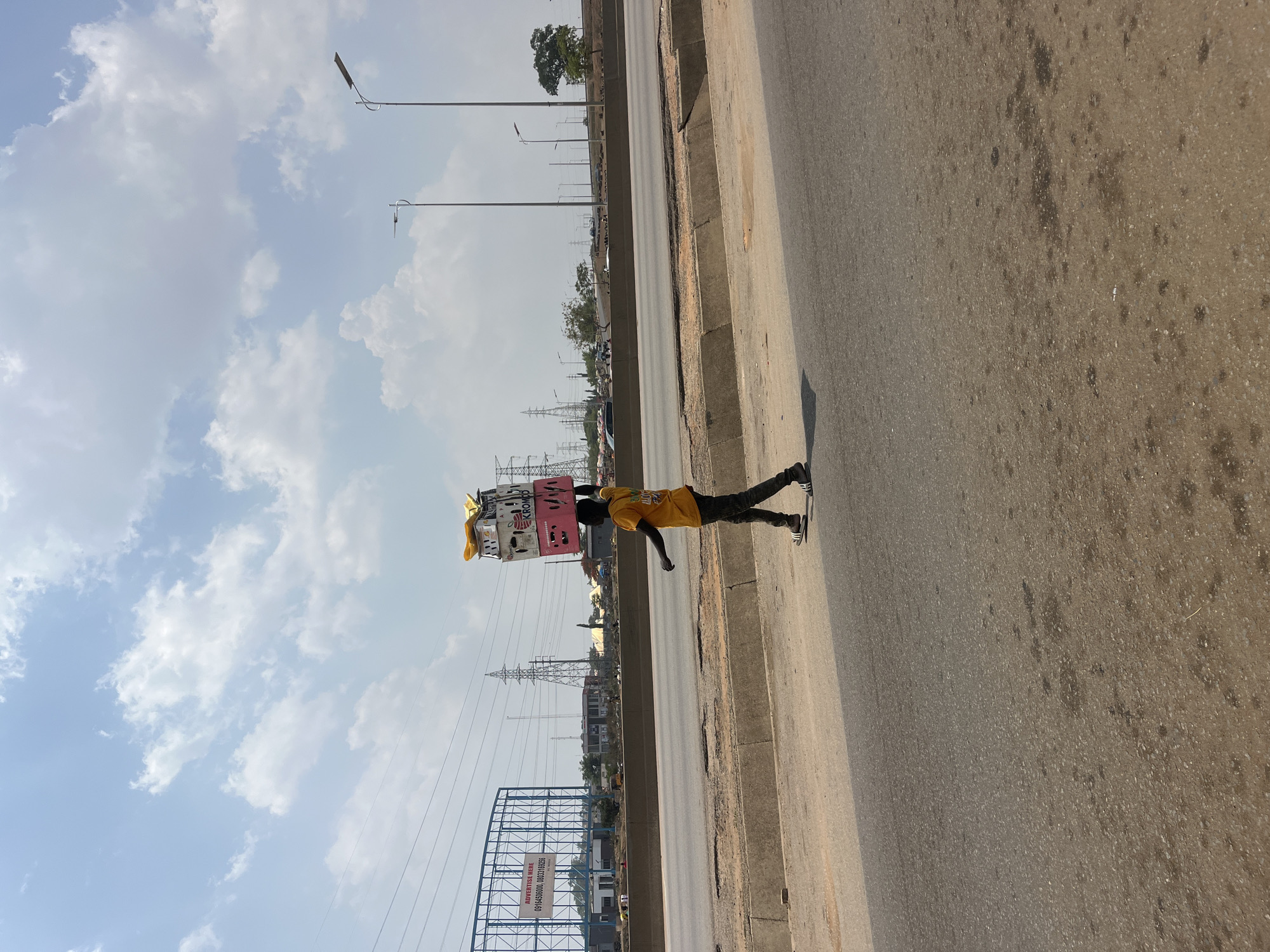 A street vendor in Galadimawa Village, Nigeria walks along the side of a paved road with an elevated sidewalk. He is carrying a stack of crates and yellow cloths on his head. The vendor is wearing a yellow shirt, dark pants, and black and white sneakers. Overhead power lines and streetlights are visible in the background, along with an advertising billboard and scattered trees. The sky is partly cloudy.