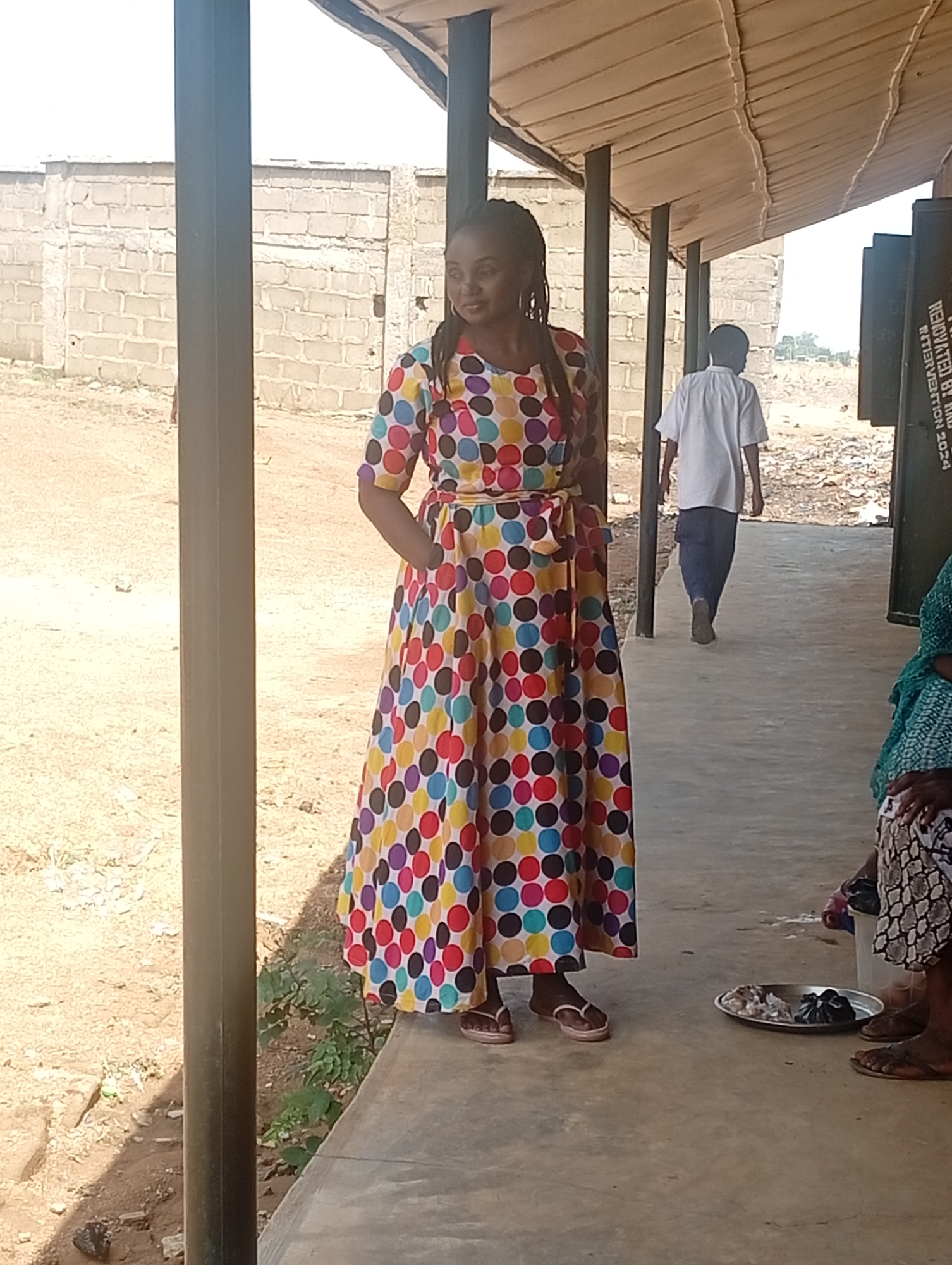 This outdoor photograph, taken in Narayi, Nigeria, captures a vibrant scene under bright daylight. The main subject is a woman standing to the left of the frame, wearing a striking ankle-length maxi dress. The dress features a lively pattern of large, multi-colored polka dots in shades of red, yellow, blue, green, brown, purple, and white on a dark background. She has her hair in dark braids and wears light-colored flip-flops. Her left hand is casually placed in a pocket, and she appears calm, looking slightly to her right.
She stands under a covered walkway, supported by sturdy dark pillars, with what appears to be a corrugated or planked roof overhead providing shade. In the mid-ground, a man in a white short-sleeved shirt and dark trousers walks away from the camera, further down the covered path. In the immediate foreground on the right, the lower legs and feet of two other individuals are visible, suggesting they are seated on the ground. A round, silver-colored tray containing small, wrapped items, possibly food or merchandise, rests near their feet.
The ground beyond the walkway is dusty, reddish-brown earth with minimal vegetation, consistent with an arid or semi-arid climate. A concrete block wall forms the background, indicating a local construction style. On a dark vertical surface in the far right background, partially visible white text reads "RENOVATED BY R..." and "INTERVENTION 2024," suggesting a renovation project. The clear, bright light indicates a sunny day, likely mid-day or afternoon. The scene conveys a sense of everyday life and commerce in a local community.
