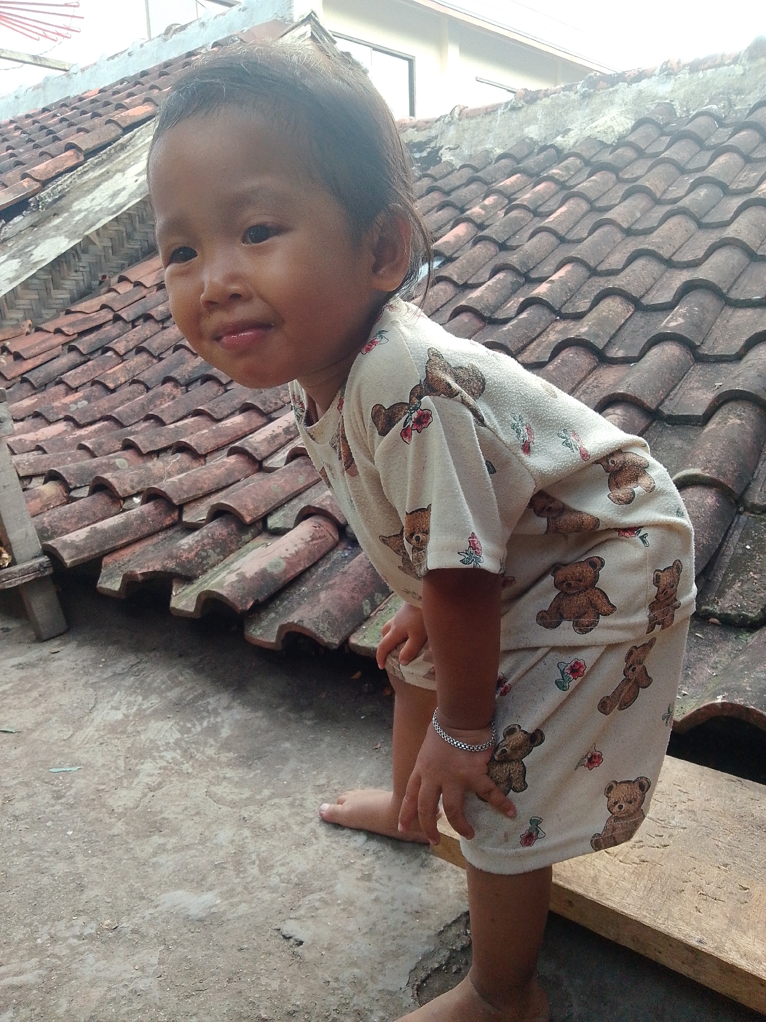 In Bandar Lampung, Indonesia, a young child is crouched on a concrete surface in front of a tiled roof. The child is wearing a light-colored onesie with a teddy bear and floral pattern. They have dark hair and are looking towards the camera with a slight smile. Their hands are visible, one resting on their leg, adorned with a silver bracelet. The tiled roof is weathered and appears to be part of an older building. The surrounding environment suggests an outdoor, possibly residential, setting.