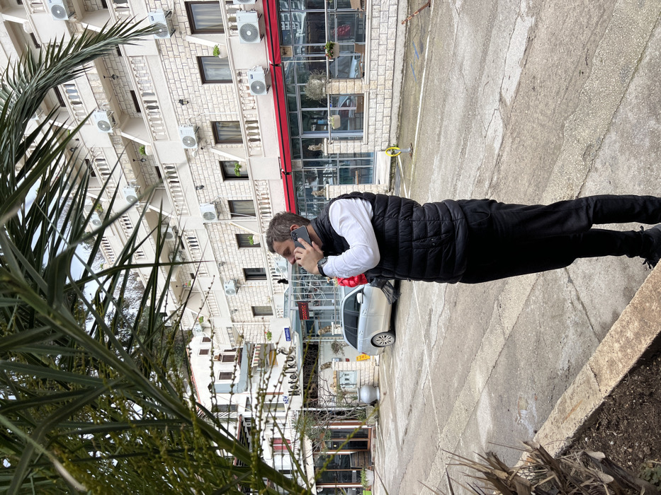 A male subject stands in an outdoor urban environment in Rafailovići, Montenegro. He is depicted in profile, facing right and looking slightly upward, holding a dark-colored smartphone to his right ear with his right hand, indicating he is engaged in a phone call. He wears a white long-sleeved shirt, a black puffer vest, black pants, and dark shoes; a watch is visible on his left wrist. A partially obscured red object, possibly a bouquet of balloons or flowers, is held in his left arm against his body. He is positioned on a gray paved surface, likely a street or parking area. In the background, a prominent multi-story building with light-colored stone cladding features numerous balconies with white balustrades and a multitude of white external air conditioning units mounted on its facade. The ground floor of this building includes commercial storefronts with red awnings and large glass windows. To the left of the subject, a sign for "Hotel Kuć" is visible, and a silver Mercedes-Benz station wagon is parked. The sky above is overcast. Palm foliage partially obscures the left and top-left foreground.