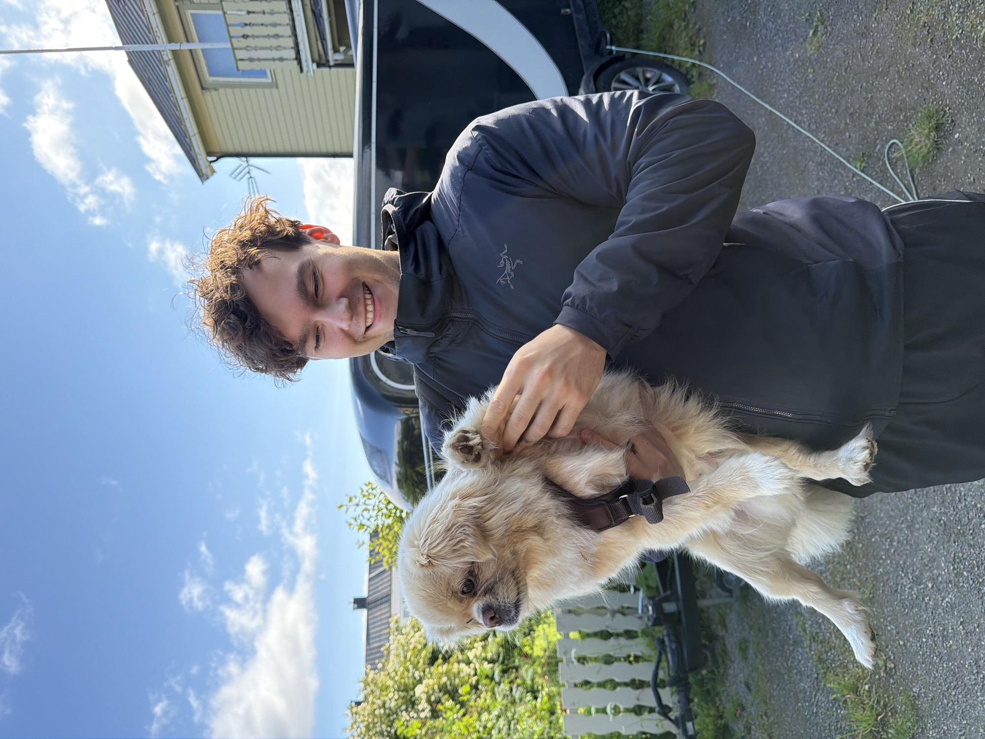 A young man with curly brown hair smiles as he holds a small, light-colored dog. The man is wearing a black hooded jacket with a logo on the left chest and black pants with a white stripe. The dog, wearing a harness, is held in his arms, and its paw is resting on his hand. Behind them, a black trailer with a white stripe is visible, as well as a light-colored house with a balcony and a white picket fence. The scene is outdoors, with a blue sky and scattered clouds overhead. The ground appears to be gravel. The photo was taken in Harstad, Norway, likely on a sunny day.
