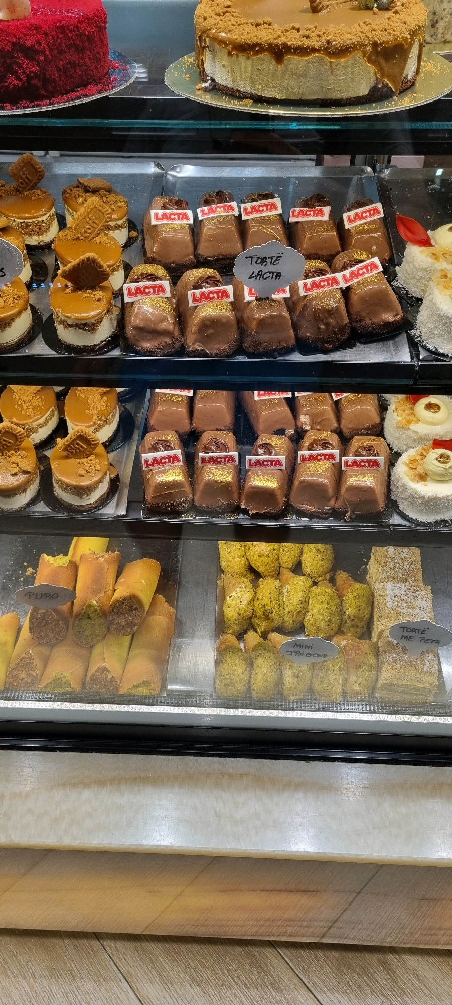 The image displays a refrigerated display case filled with various desserts. In the top section, two large cakes are visible: one is a deep red cake, and the other is a caramel-colored cake with a crumb topping. Below these, several rows of individual desserts are arranged. The middle shelf contains rows of small, chocolate-covered cakes, many of which have "LACTA" labels and some are sprinkled with gold edible glitter. To the right of these chocolate cakes are two white cakes decorated with shredded coconut and candied fruit. The bottom shelf features several types of pastries. On the left are cylindrical pastries filled with a dark mixture and partially covered in a green coating, with a "PURO" label. To the right of these are small, cone-shaped pastries coated in a green substance, some with "MINI" labels. Further to the right on this shelf are rectangular layered cakes with a light brown topping. The overall environment appears to be a bakery or patisserie. The location is identified as being in the city of Bashkia Durrës, Albania. There are no people visible in the image, and the focus is solely on the presented food items.