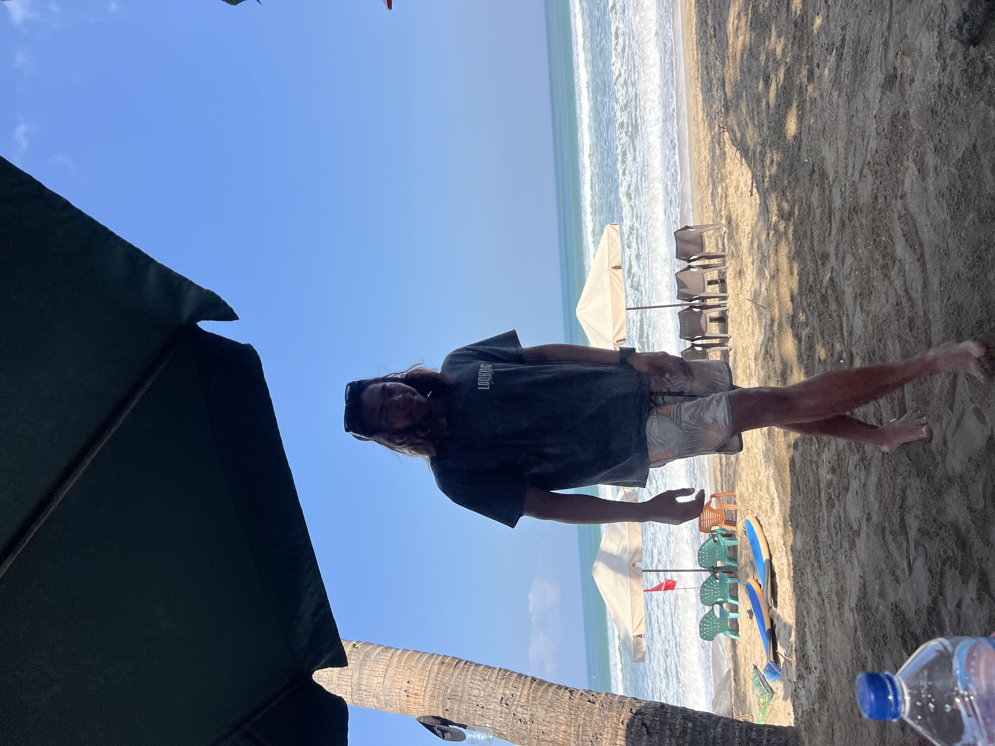 An outdoor shot captures a man standing on a sandy beach in Kuta, Indonesia, with the ocean and sky as the backdrop. The man is wearing a dark t-shirt and patterned shorts, with sunglasses perched on his head. He stands partially under a large, dark green umbrella, with a palm tree to his left. In the mid-ground, beach chairs and umbrellas are set up, and two surfboards are lying on the sand. The ocean waves are visible in the background, and the sky is a clear blue. A plastic water bottle is in the foreground, on the left side of the frame. The lighting suggests it is daytime, with bright sunlight casting shadows on the sand.
