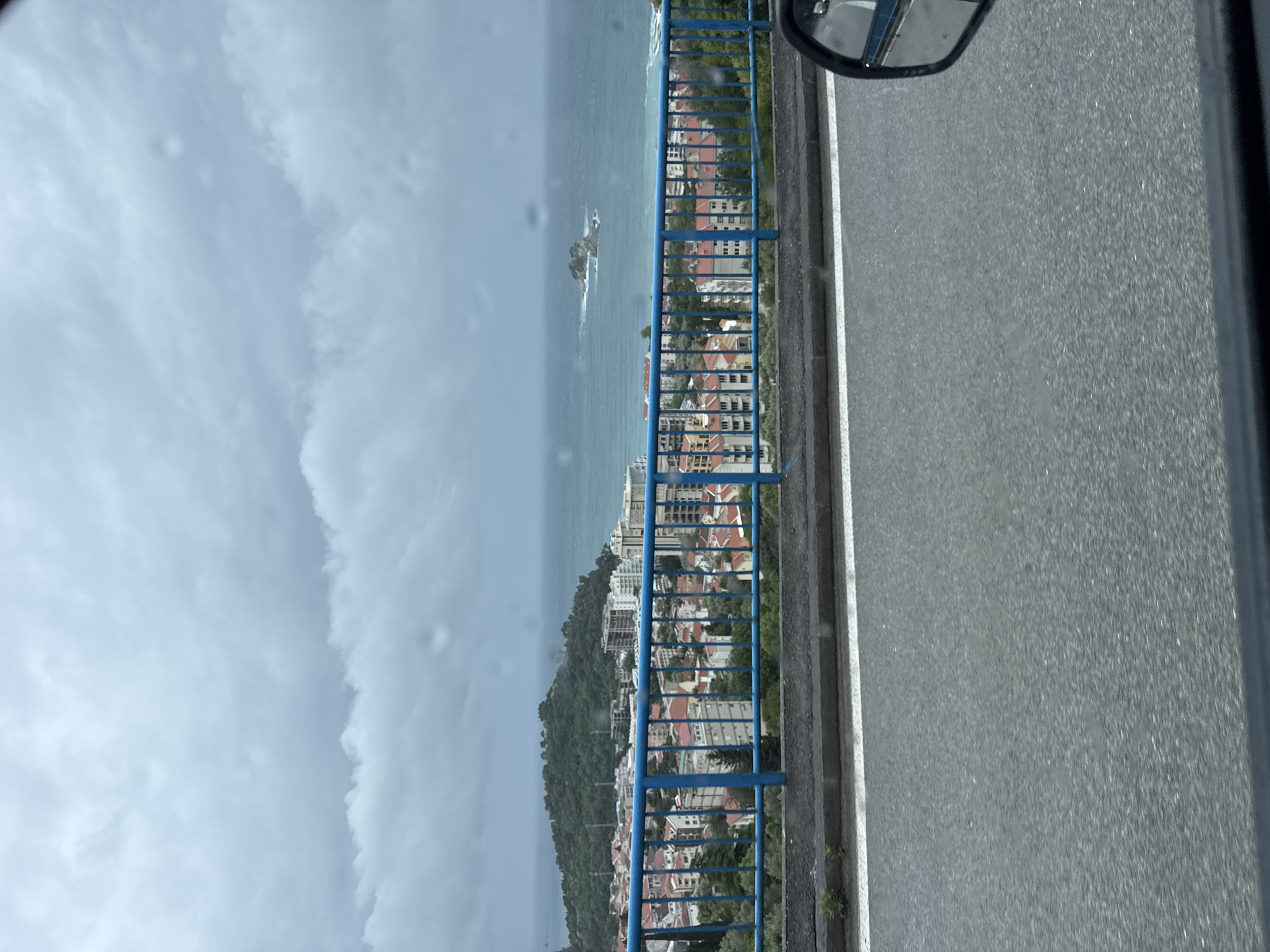 The image shows a view from a bridge in or near Stupovi, Montenegro. The Adriatic Sea is visible with a small rocky island. Coastal buildings with red-tiled roofs are interspersed with greenery on a hillside. The blue bridge railing runs along the side of the road, visible in the foreground. A car side mirror is present in the upper right corner. The sky is overcast with grey clouds.