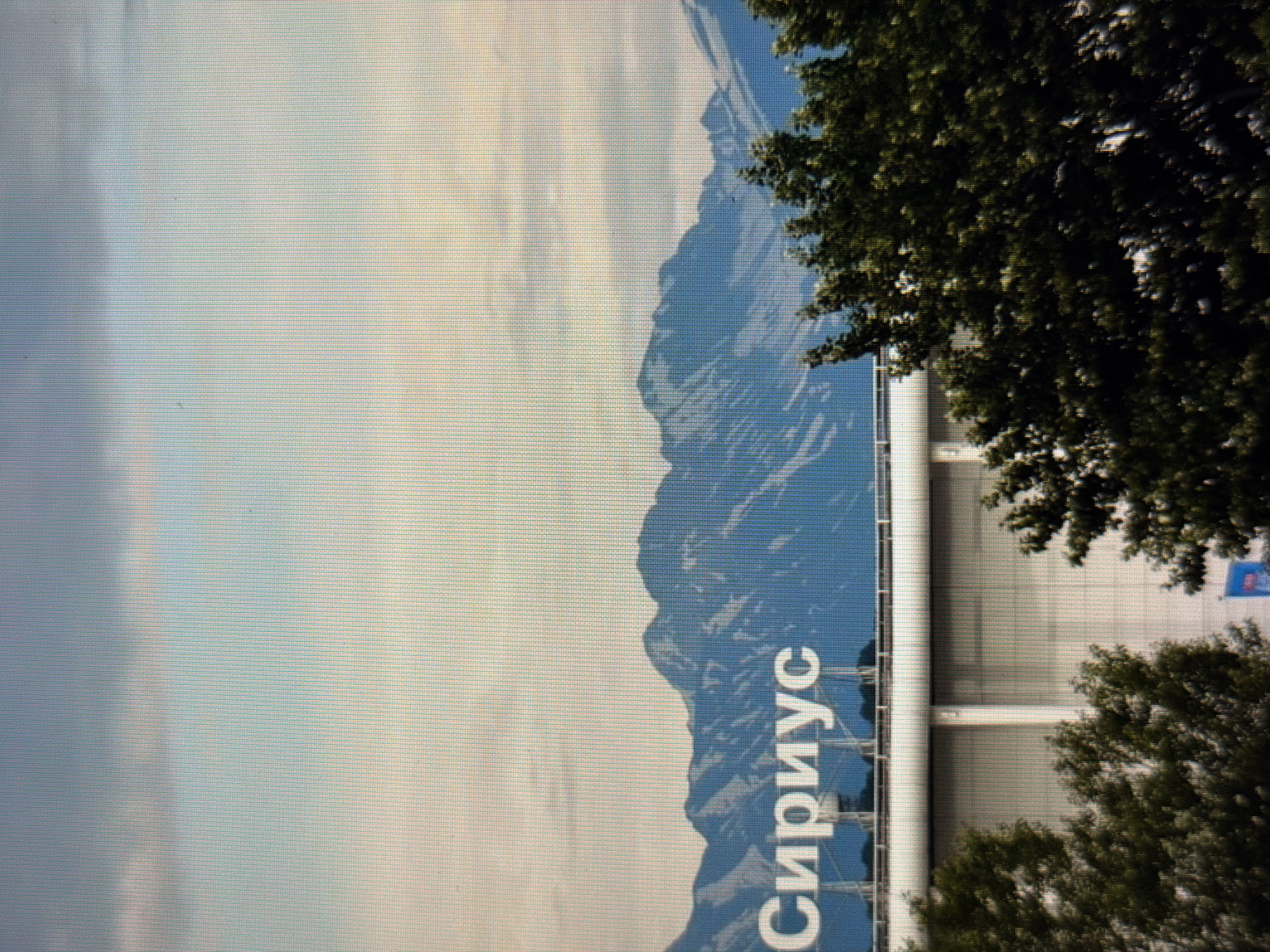 This is a photograph taken from a low angle, looking up at a building. The building appears to be quite large, with a white facade and vertical white columns. A large blue mural or banner is visible on the side of the building, depicting a mountainous landscape. Text in white Cyrillic characters, "СИРИУС," is prominently displayed on the blue mural, oriented vertically. The word "Сириус" translates to "Sirius." Green trees with dense foliage are in the foreground and to the right of the building, partially obscuring the view. The sky is visible in the upper left portion of the image, with light clouds and a soft, possibly diffused daylight. The overall impression is that of a modern building in a setting with natural elements. The orientation of the image suggests it might have been taken with the camera held sideways. The image appears to have been captured on a screen, as it shows a pixelated grid pattern.