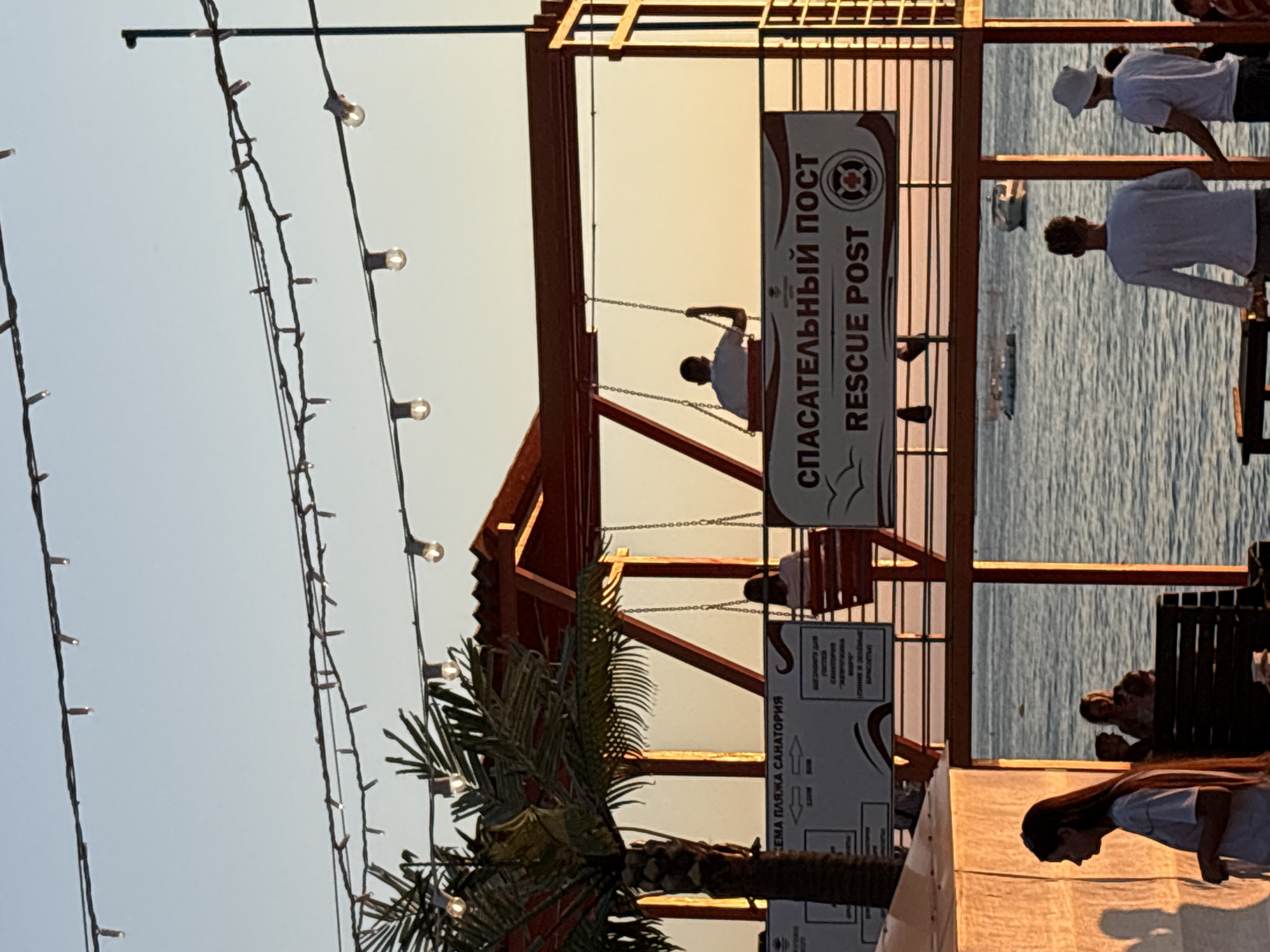 This is a wide shot taken at sunset of a rescue post building located on a beach, likely in Novorossiysk, Russia, given the Cyrillic text on the signage. The building, painted orange and red, has a raised platform with swings. On one swing, a person is seated, facing away from the camera. Another person is on a different swing, also facing away. A sign on the building reads "СПАСАТЕЛЬНЫЙ ПОСТ RESCUE POST" in Cyrillic and English, with a lifebuoy symbol. To the left of the rescue post is a palm tree. The background shows the calm sea with a few boats visible, and the sky is a gradient of blue and orange due to the sunset. In the foreground, several people are visible. On the left, a woman with long hair is seen in profile. To the right, two men, one wearing a white hat, stand facing the sea. The overall impression is a relaxed, late-day scene at a seaside resort.