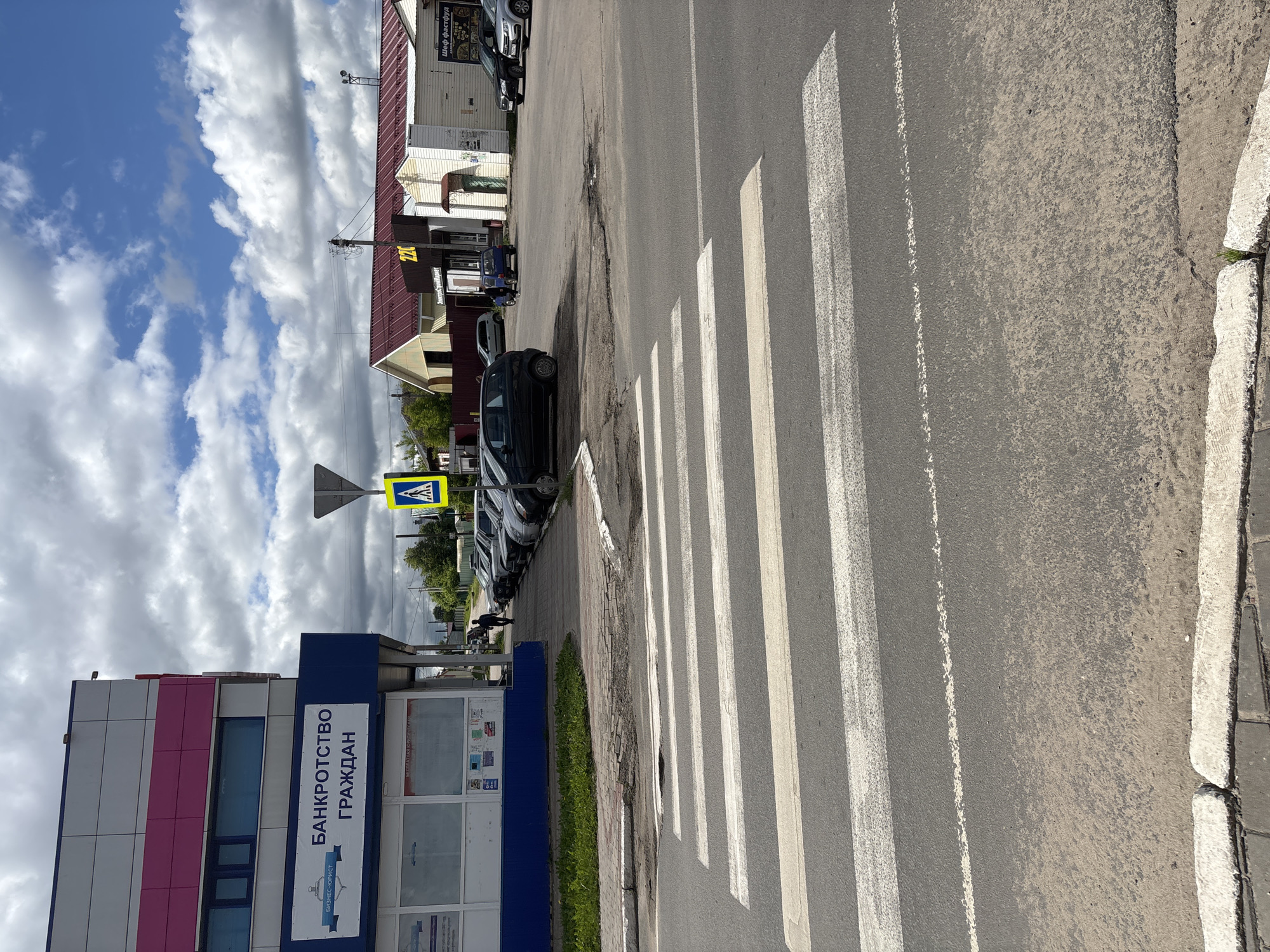 The image captures a street scene in Красные Баки, Russia, under a bright, partly cloudy sky. The photograph is taken from a slightly elevated perspective, looking across an asphalt road.
In the foreground, a prominent pedestrian crossing, marked by wide white "zebra" stripes, stretches across the road. The asphalt surface of the road shows signs of wear. To the right of the road, a concrete curb separates it from a paved sidewalk.
Along the left side of the road, several cars are parked bumper-to-bumper. Beyond the parked vehicles, there is a row of buildings. The most prominent building in the immediate left foreground features a modern facade with panels of pink and white on its upper section and a dark blue base. A sign on its blue section clearly displays the Russian text "БАНКРОТСТВО ГРАЖДАН" (Bankruptcy of Citizens) and "БИЗНЕС-ЮРИСТ" (Business Lawyer).
Further down the street, other buildings are visible, including one with a reddish-brown roof and a sign in Russian that reads "Шеф фастфуд" (Chef Fast Food), along with a yellow sign displaying "24" and what appears to be part of "час" (hour), likely indicating 24-hour service. Another building has light-colored siding. A single person is visible walking on the sidewalk in the distance, near the parked cars.
Notable details include a yellow diamond-shaped road sign with a blue square and white pedestrian symbol, indicating a pedestrian crossing ahead. The sky is bright blue with numerous fluffy white clouds, suggesting good weather. Strong shadows cast by the sun indicate it is daytime, likely mid-morning or afternoon.