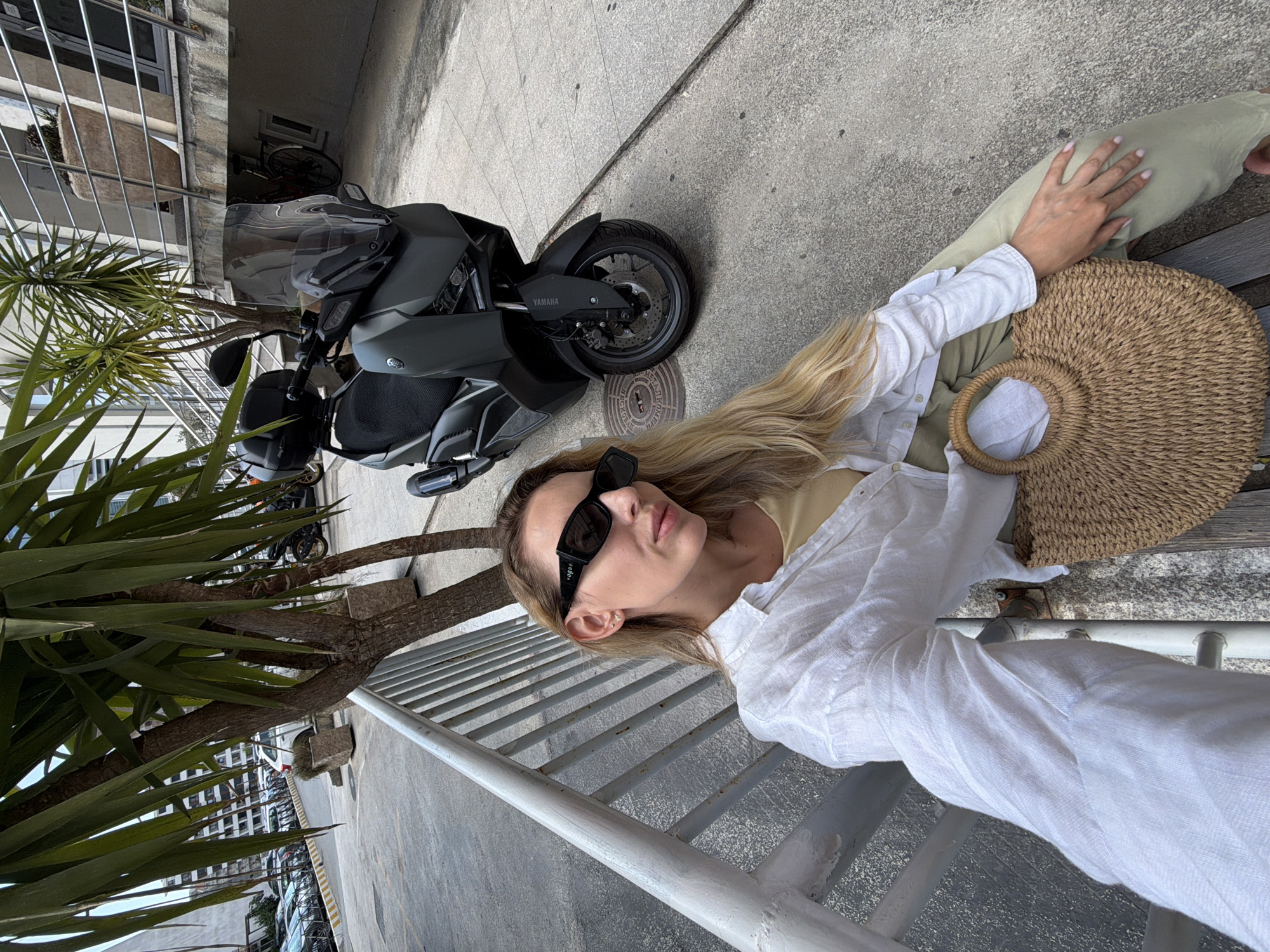A woman with blonde hair, wearing sunglasses, a white unbuttoned shirt over a tan top, and olive green pants, sits on a bench next to a metal railing. She is holding a straw bag on her lap. Behind her, a dark grey Yamaha scooter is parked on a paved surface. The scene appears to be on a street in Budva, Montenegro, with buildings and parked cars visible in the background.
