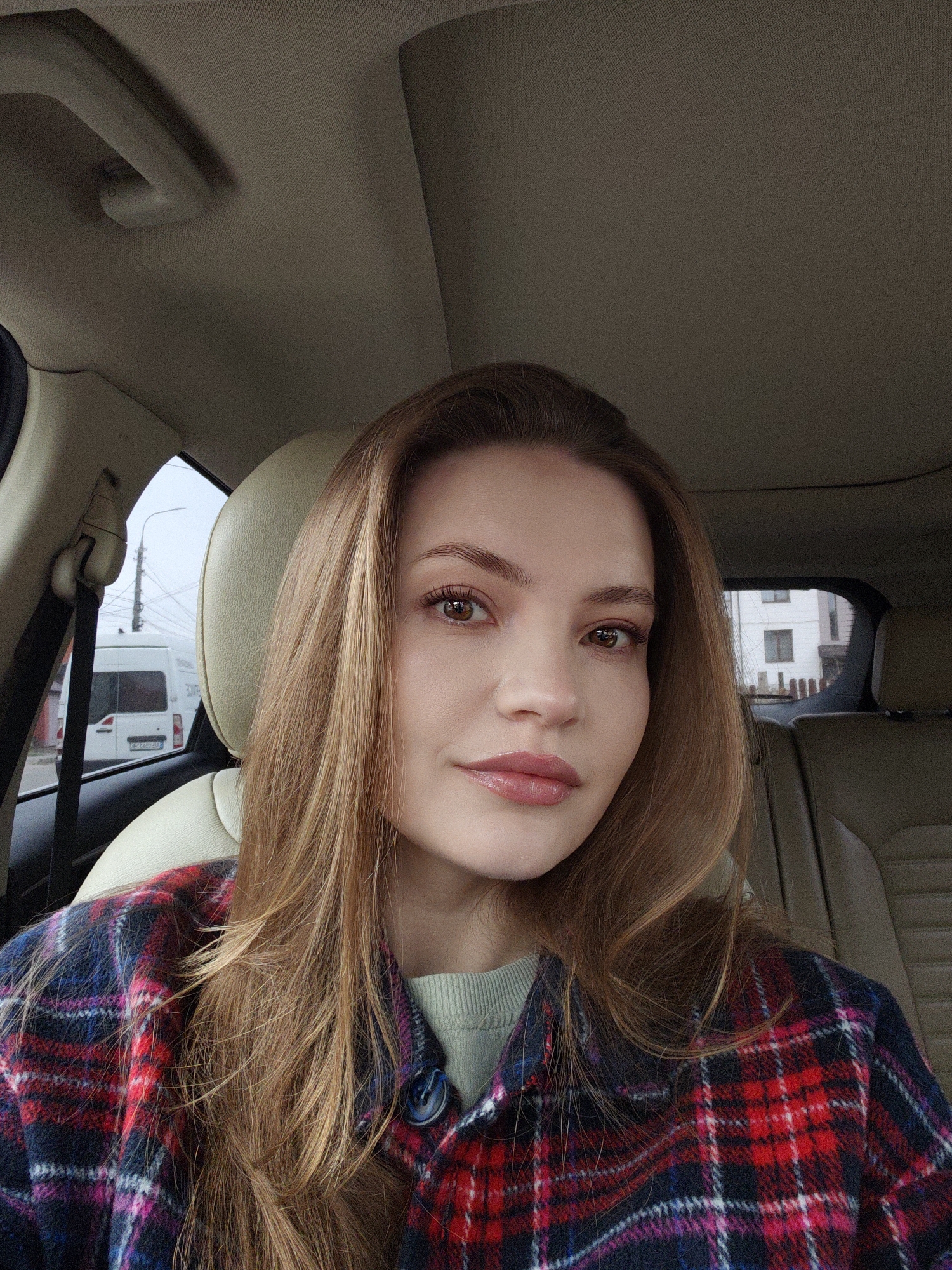 The image shows a woman in the front seat of a car. She is fair-skinned with light brown hair and brown eyes. She wears a red and blue plaid jacket over a light green sweater. The interior of the car is beige, with a headrest visible behind her. Outside the car window, a white van is visible, and a building and power lines are in the background. Based on the license plate on the van and surrounding architecture, the location is Vinnytsia, Ukraine. The woman is looking directly at the camera, seemingly taking a selfie.