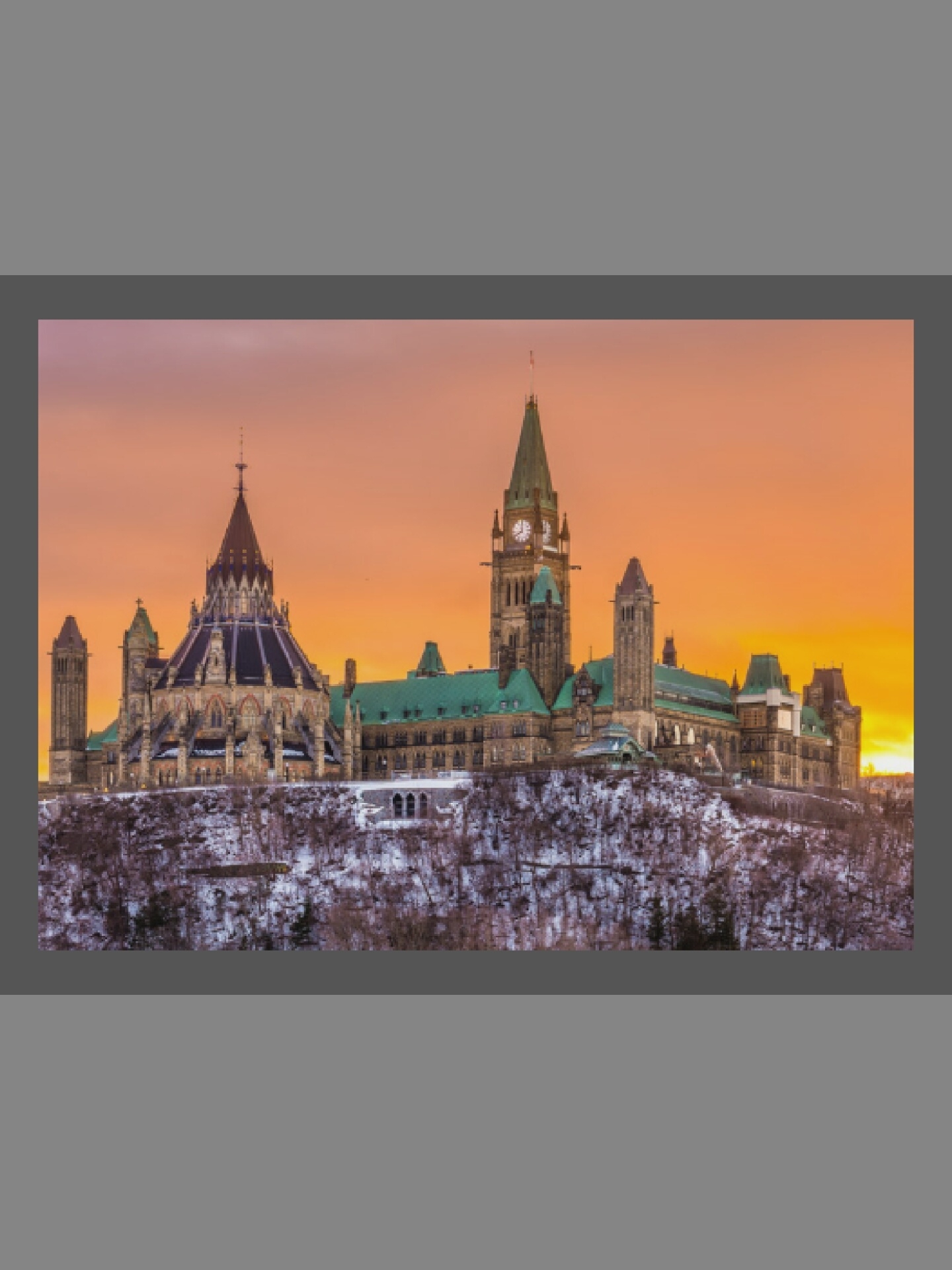 The image captures a majestic view of the Parliament of Canada buildings in Ottawa, Canada, set against a stunning sunset or sunrise sky. The main subjects are the Gothic Revival parliamentary complex, including the distinctive Peace Tower with its clock and the domed Library of Parliament to the left. The buildings are perched atop a snow-covered, treed hillside, suggesting a winter scene.
The sky is a dramatic blend of vibrant orange, yellow, and soft pink hues, creating a warm glow that contrasts with the cool tones of the snow and the detailed stone architecture. The roofs of the buildings are green, possibly copper, adding another color element to the scene. No specific human activity is visible, making the monumental architecture and the natural beauty of the winter landscape the focal point. The image conveys a sense of grandeur and tranquility. There is no visible text in the scene.
