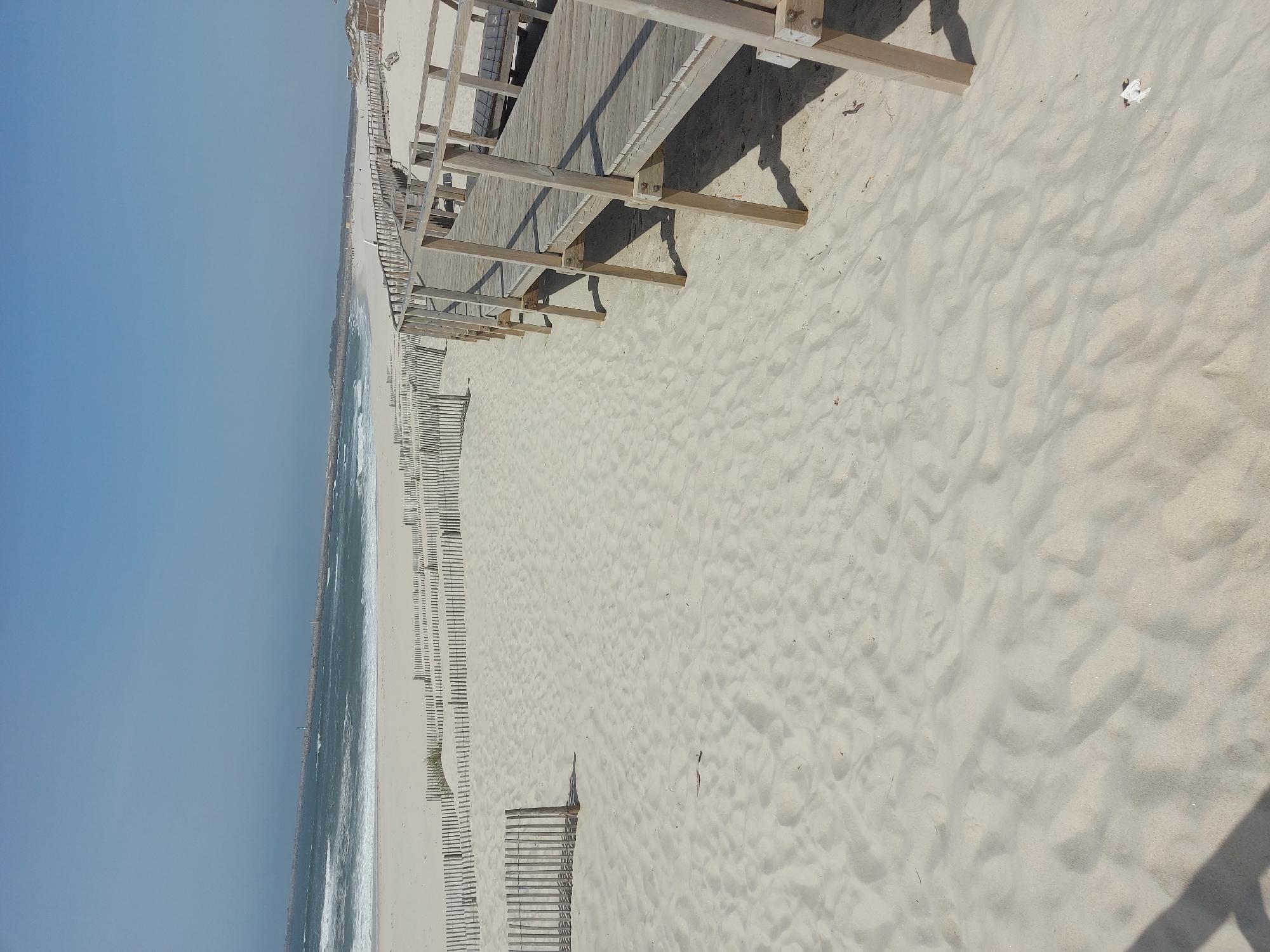 This photograph shows a view of a beach in Gafanha da Nazaré, Portugal. The scene includes a sandy beach, an ocean with waves, a walkway constructed of wood, and numerous wooden fences placed along the beach. The sky is clear and blue. The photograph is taken from an elevated position. The sand is beige and shows patterns of wind and foot traffic. The wooden fences run along the beach parallel to the ocean, and the walkway is positioned higher, casting shadows on the sand. There are no people visible in the image.